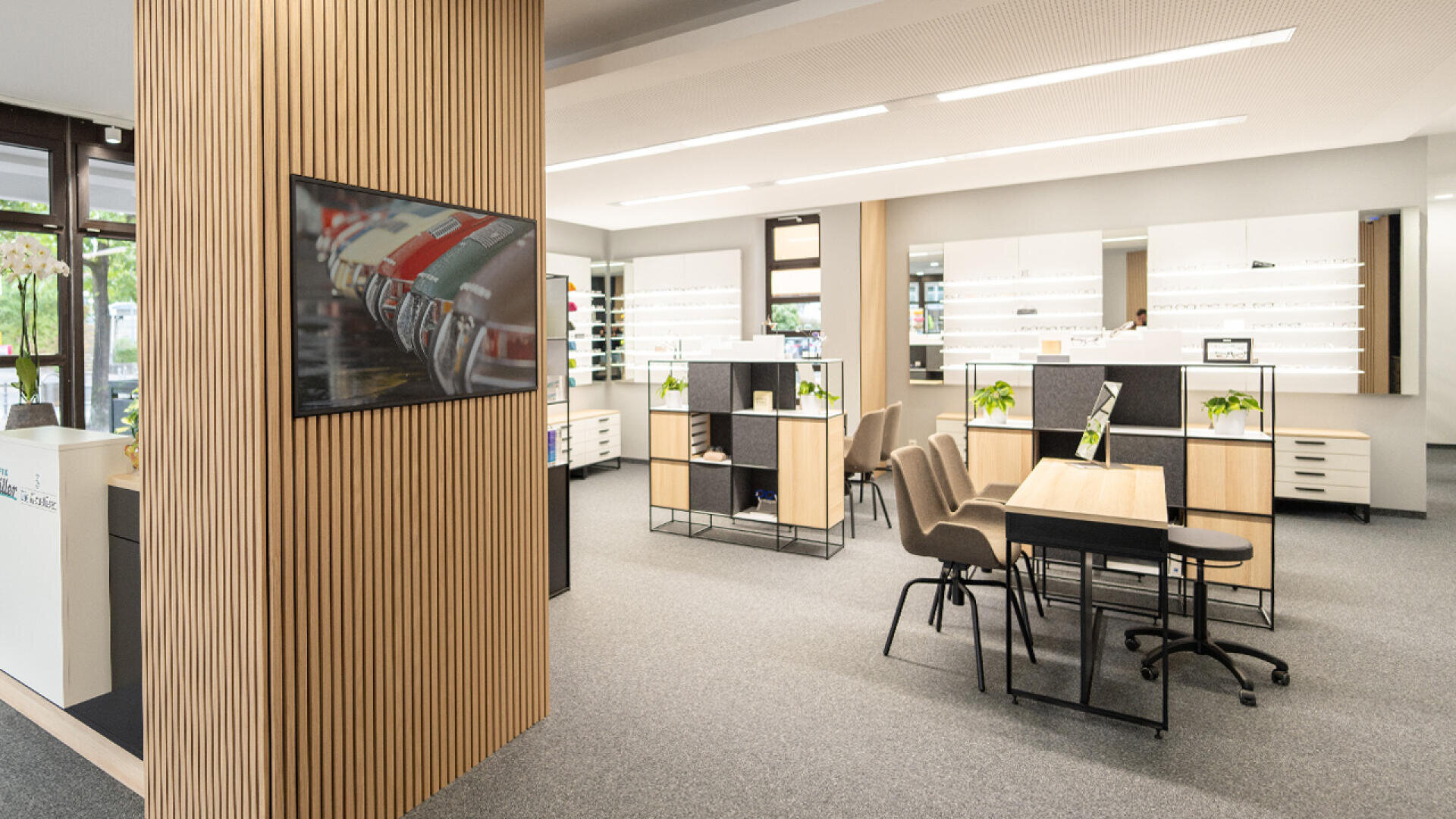 A modern, well-lit optician’s office with display shelves of eyeglasses, wooden accents, a meeting desk with chairs, potted plants, and a TV on the wall. Large windows let in natural light.