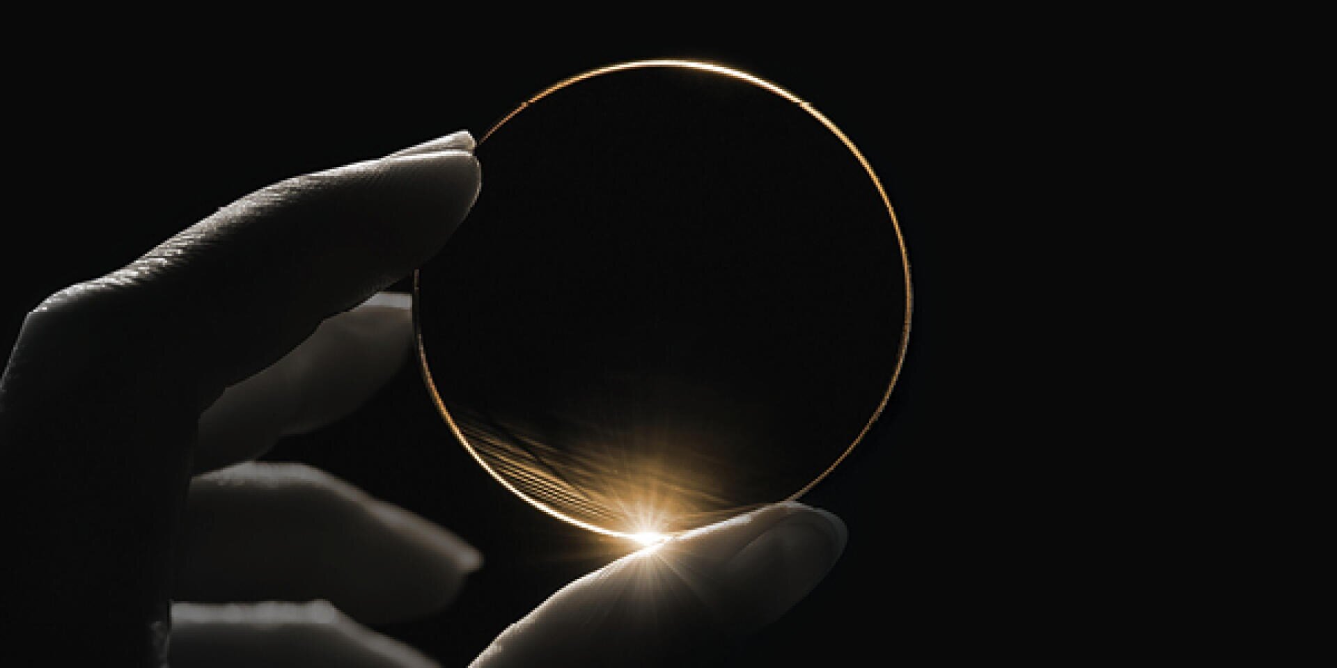 A hand wearing a glove holds a round, transparent lens or pane of glass in front of a dark background, with the light illuminating the edge and creating a bright, luminous rim.