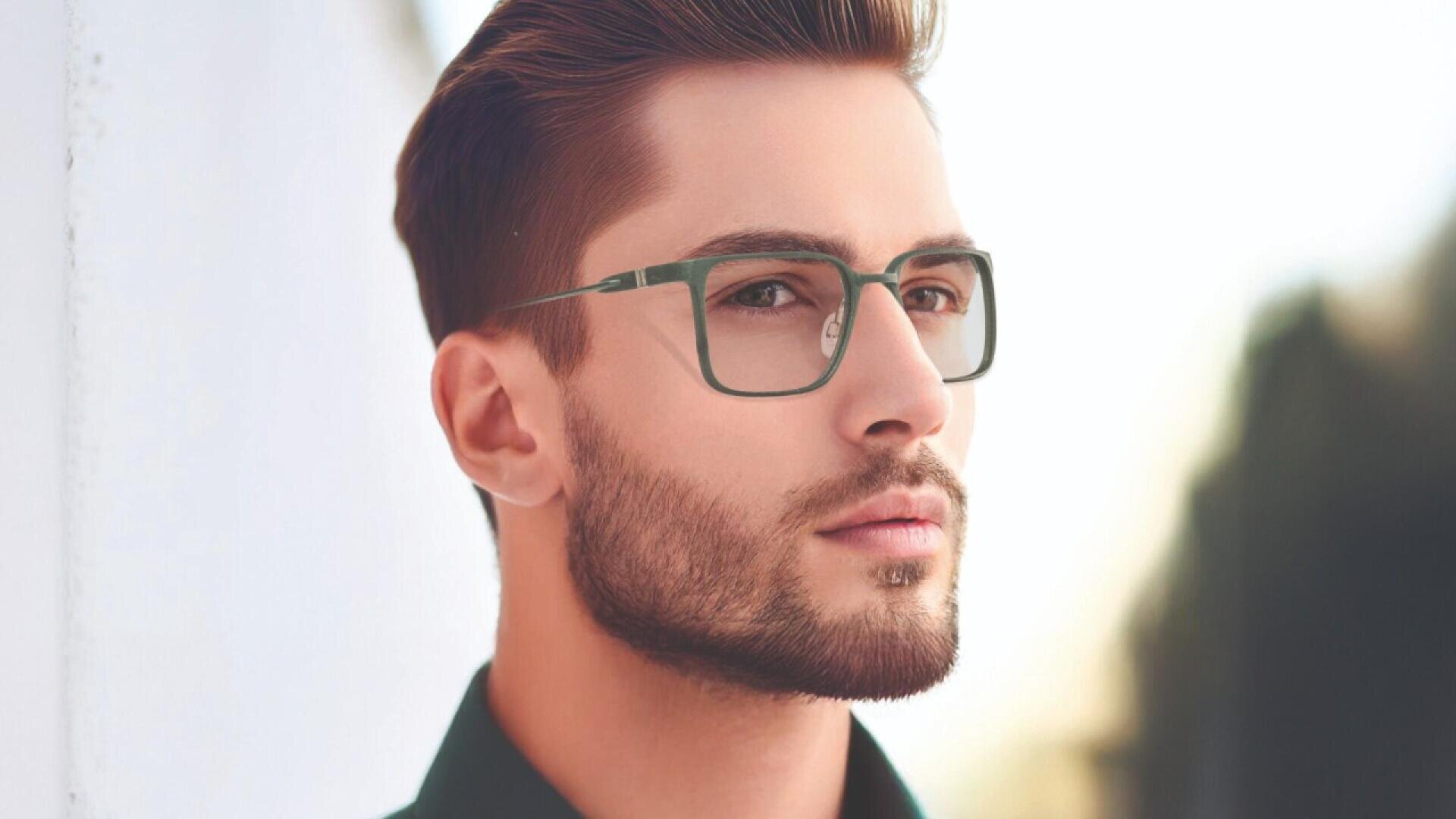 A young man with short brown hair, a trimmed beard, and wearing green-rimmed glasses looks thoughtfully into the distance, standing outdoors near a white wall.