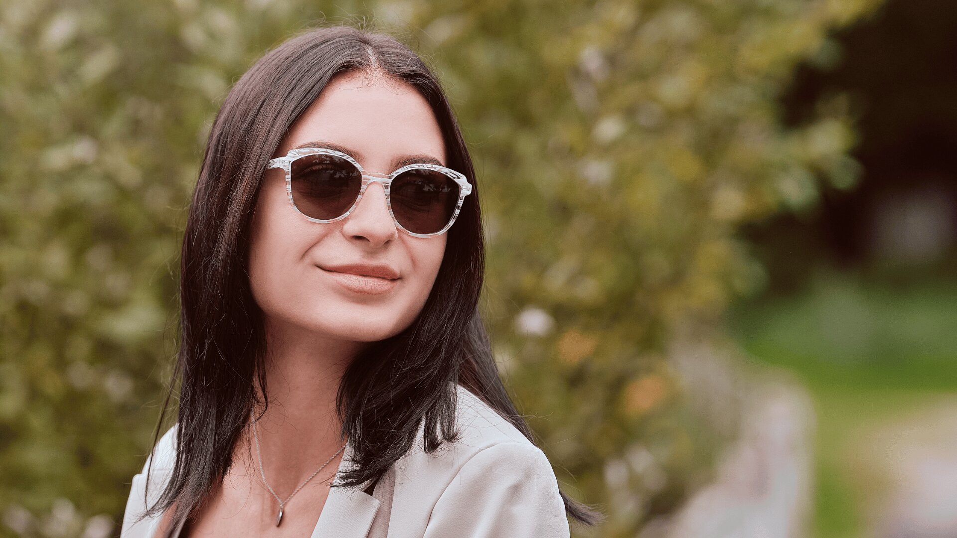 Eine Frau mit langen dunklen Haaren, die eine runde Sonnenbrille und einen hellen Blazer trägt, lächelt leicht, während sie im Freien steht und das Grün im Hintergrund sieht.