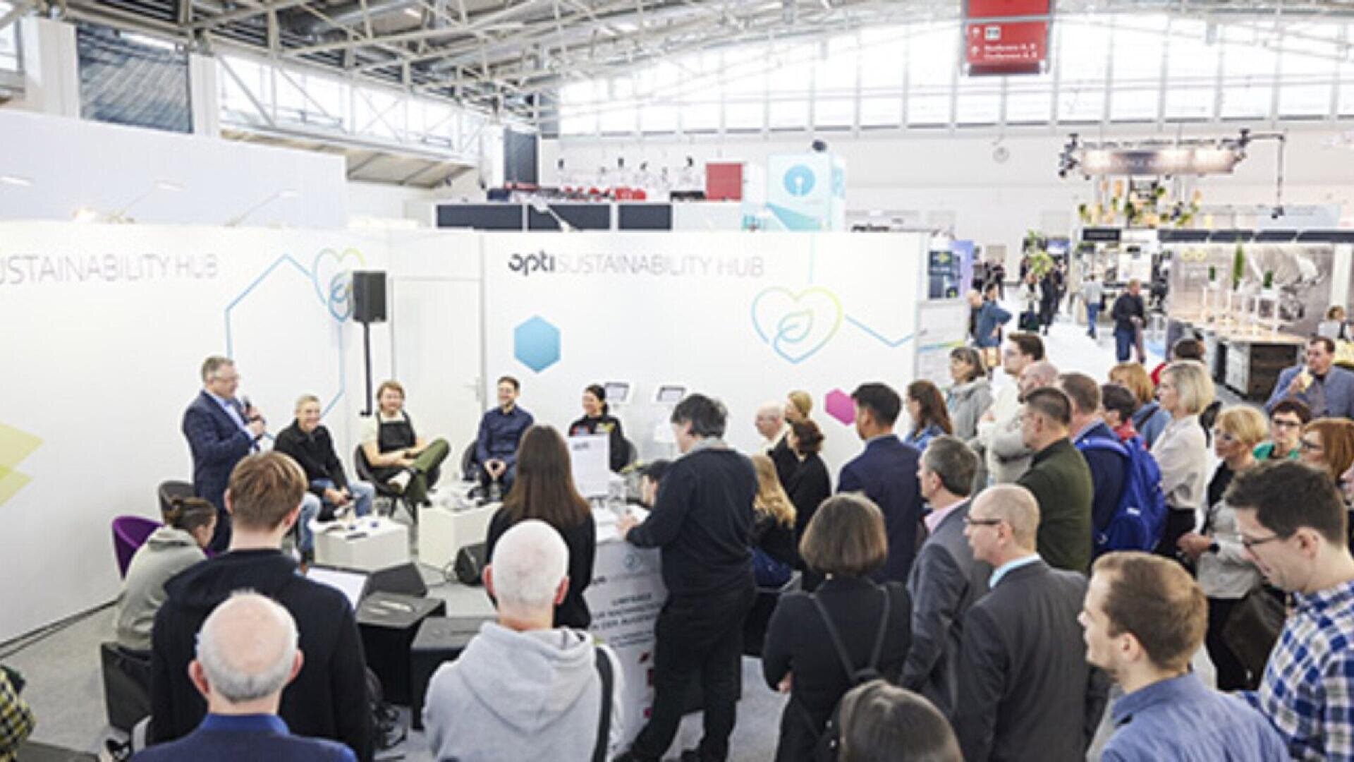A group of people listens to a panel discussion at a sustainability event in a large, bright exhibition hall with informational displays and signage in the background.