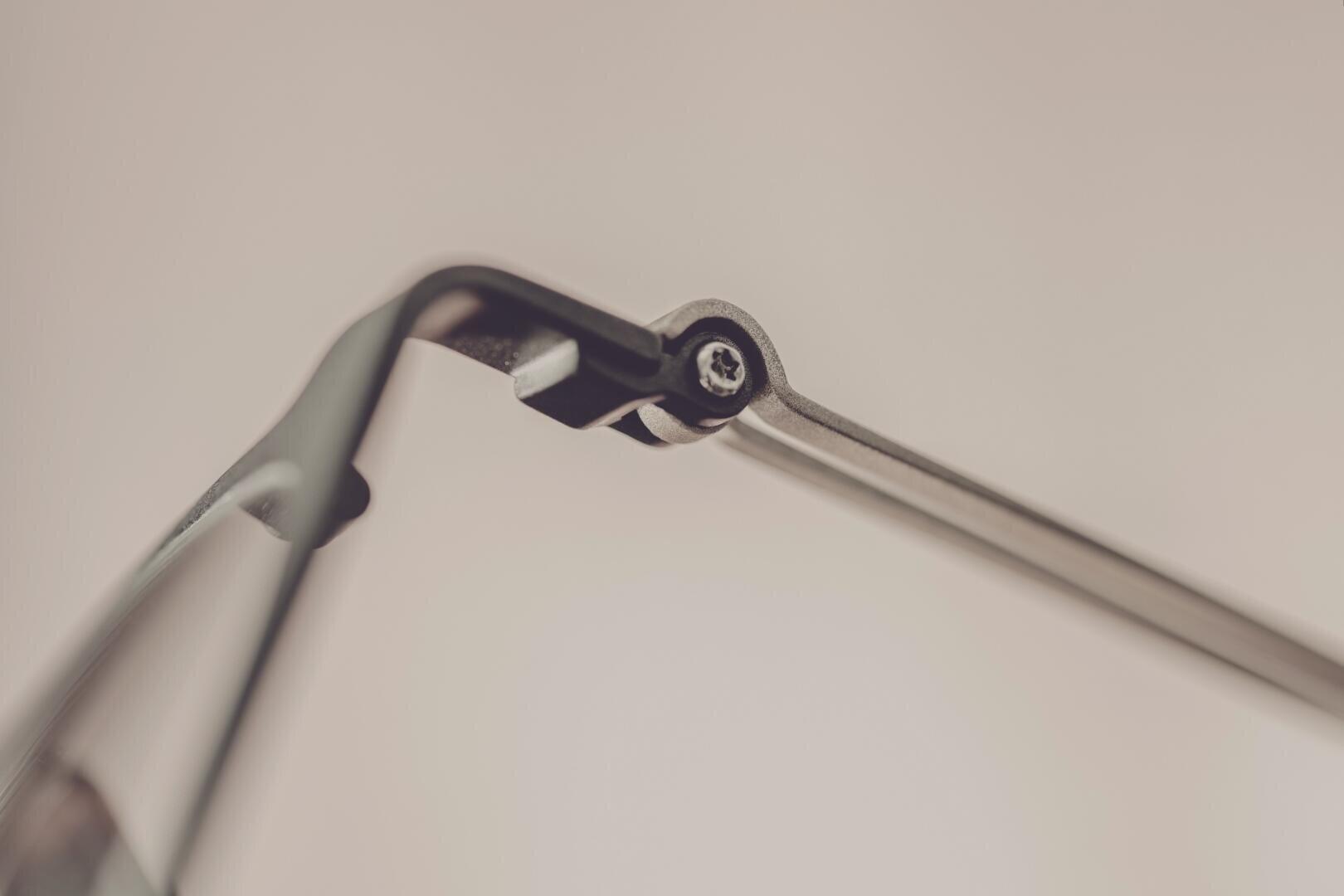 Close-up of the hinge on the arm of a pair of eyeglasses, showing the screw and metal construction with a blurred, neutral background.