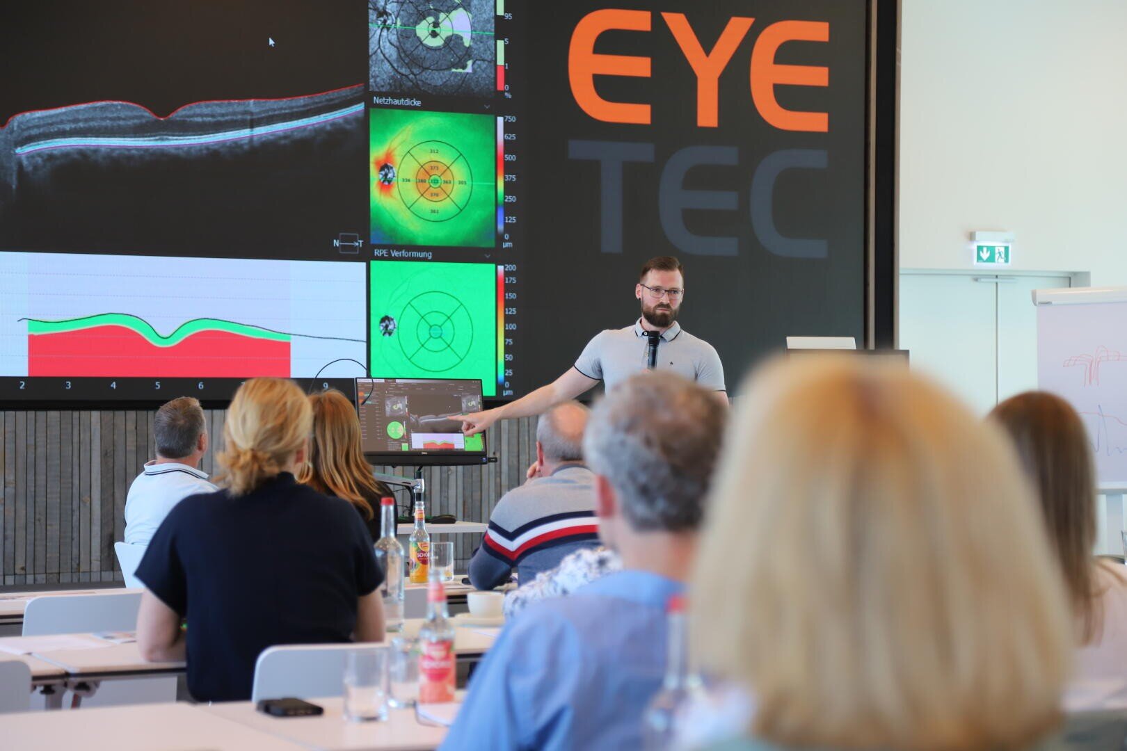 Ein Mann hält vor einem Publikum einen Vortrag über Augentechnologie, während hinter ihm auf einem großen Bildschirm Augenscans und Diagramme angezeigt werden. Das Wort EYE TEC ist deutlich an der Wand zu lesen.