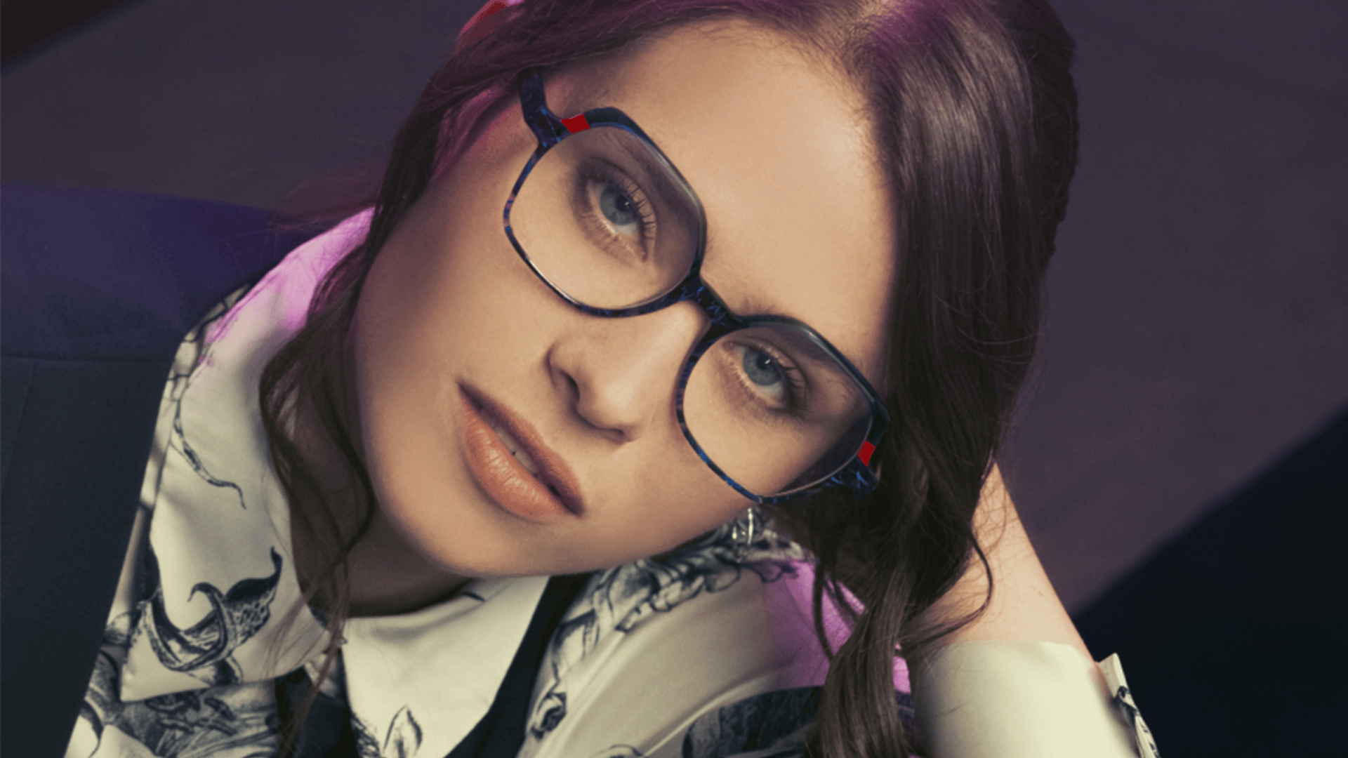 A person with long brown hair and glasses looks up at the camera. They are wearing a white shirt with a black pattern and a dark vest. The background is blurred and softly lit.