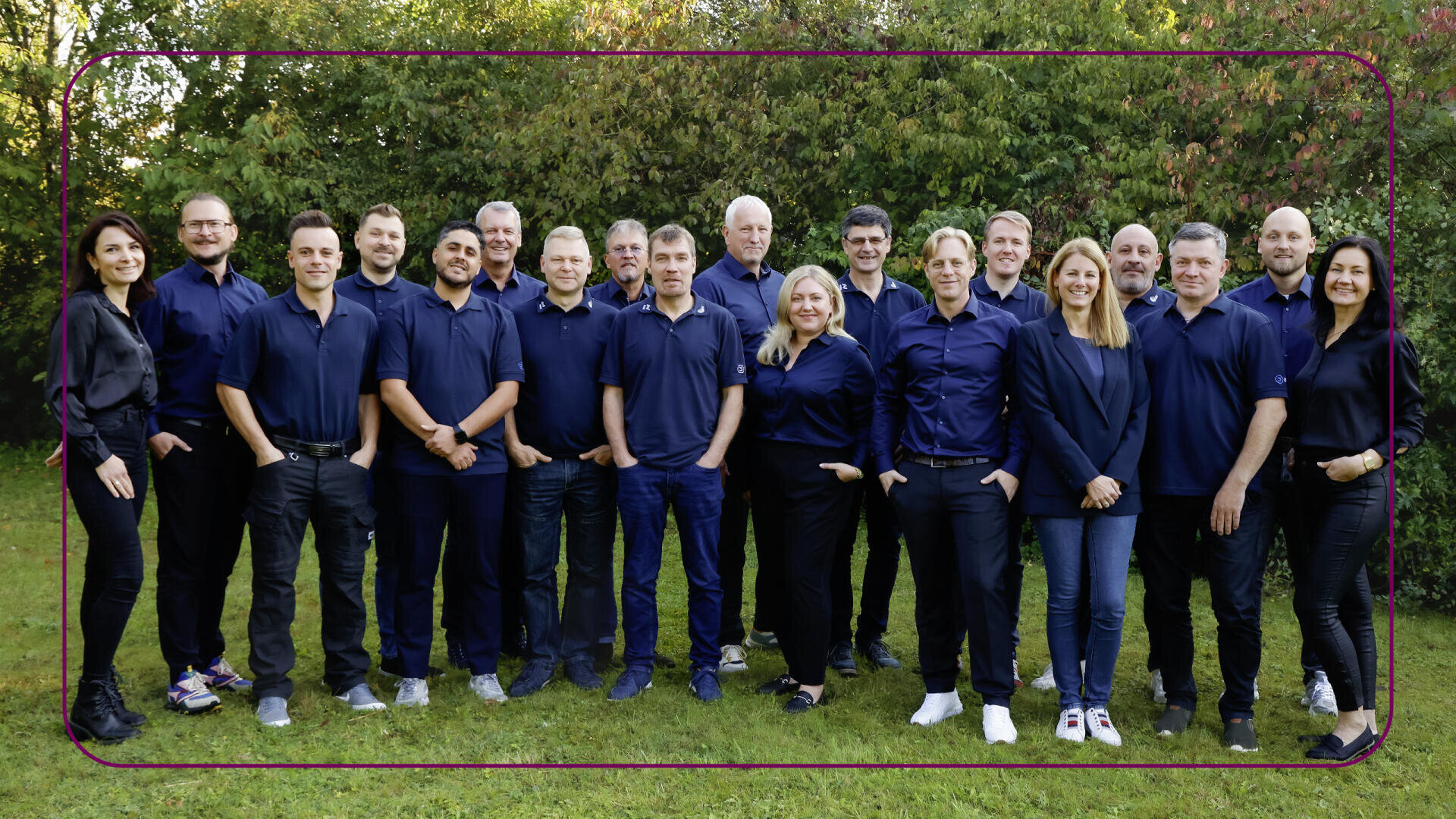 Eine Gruppe von achtzehn Erwachsenen, meist in dunkelblauen Hemden und Freizeithosen, steht im Freien auf einer Wiese mit grünen Bäumen im Hintergrund und lächelt in die Kamera.