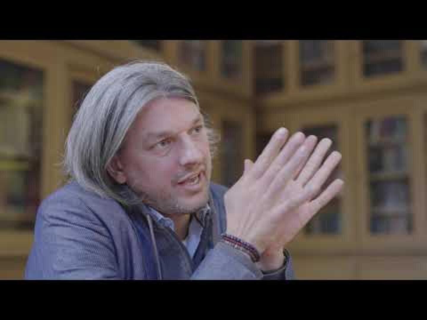 A man with shoulder-length gray hair and a short beard, wearing a blue jacket, sits in an interior room in front of a bookshelf and gestures with his hands as he speaks.