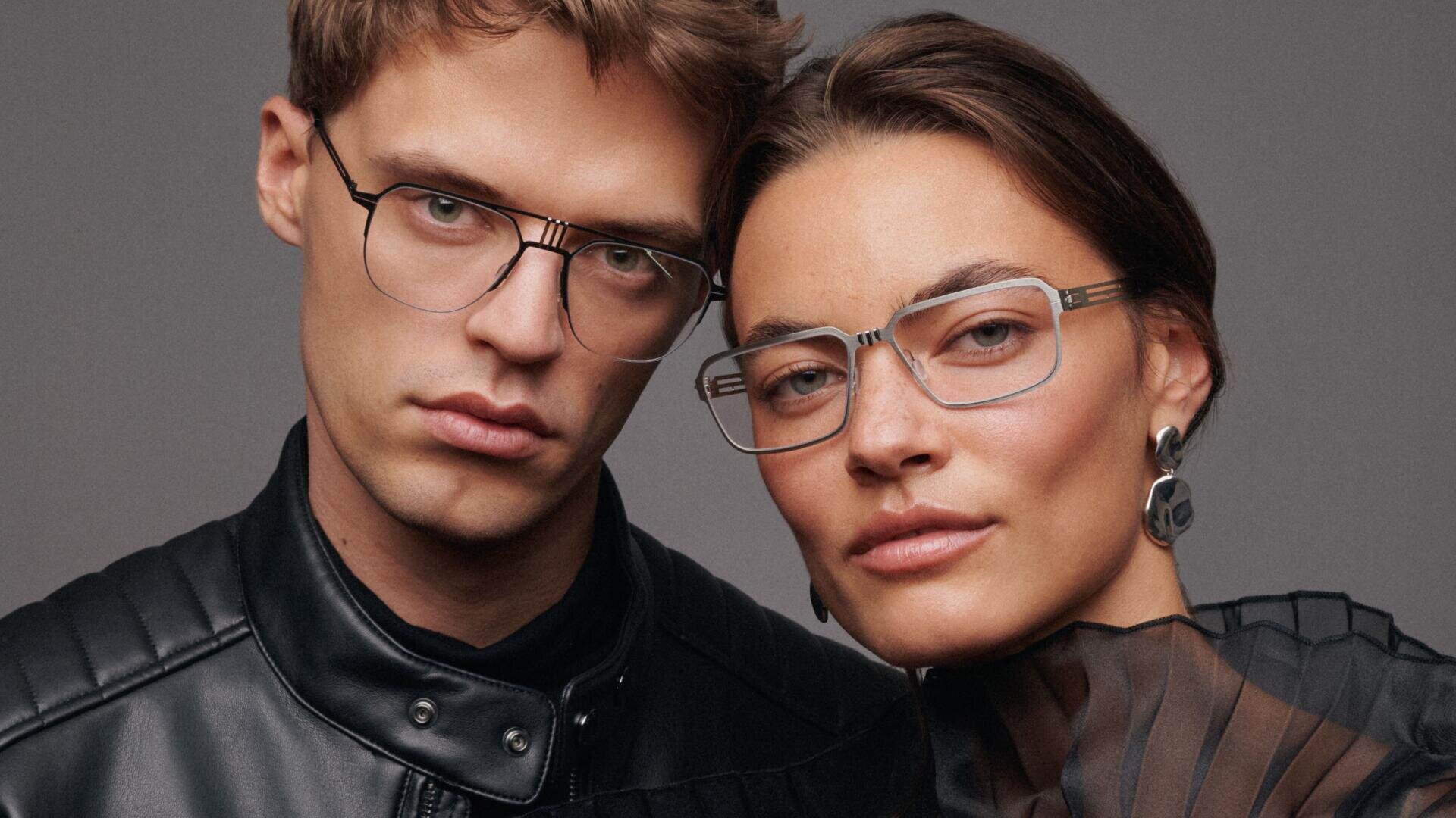 A man and a woman, wearing elegant glasses and modern, dark clothing, pose close together in front of a plain gray background. Both have a neutral expression and are looking directly into the camera.