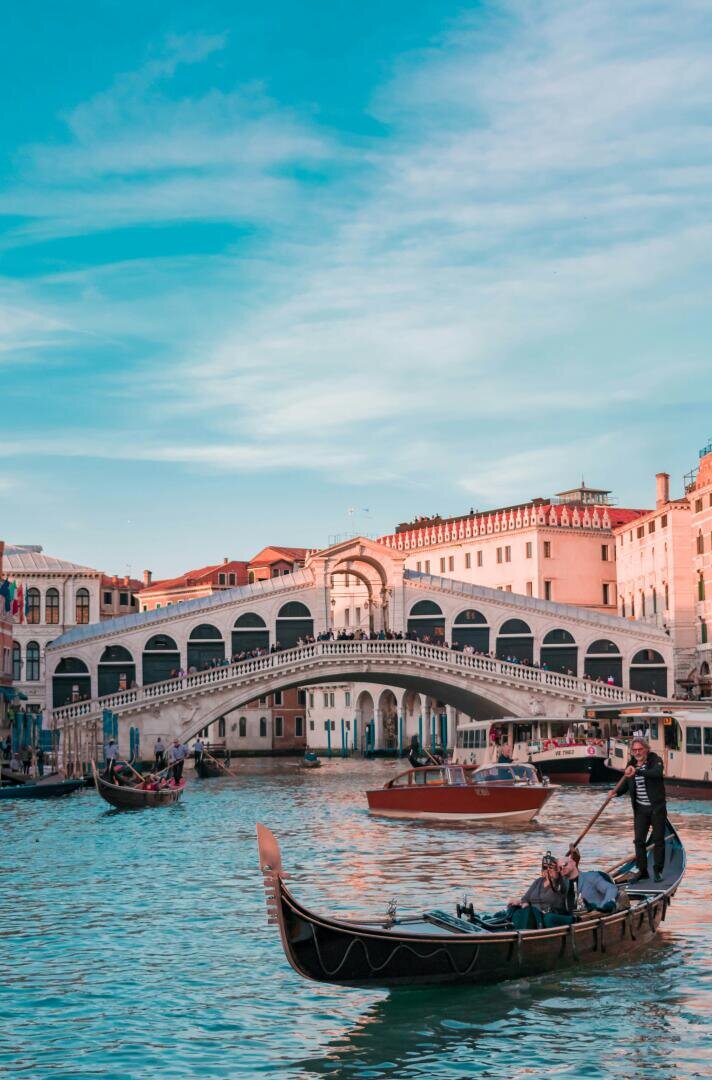 A gondolier steers a gondola with passengers through a canal in Venice, with the historic Rialto Bridge and the surrounding buildings under a blue sky in the background.