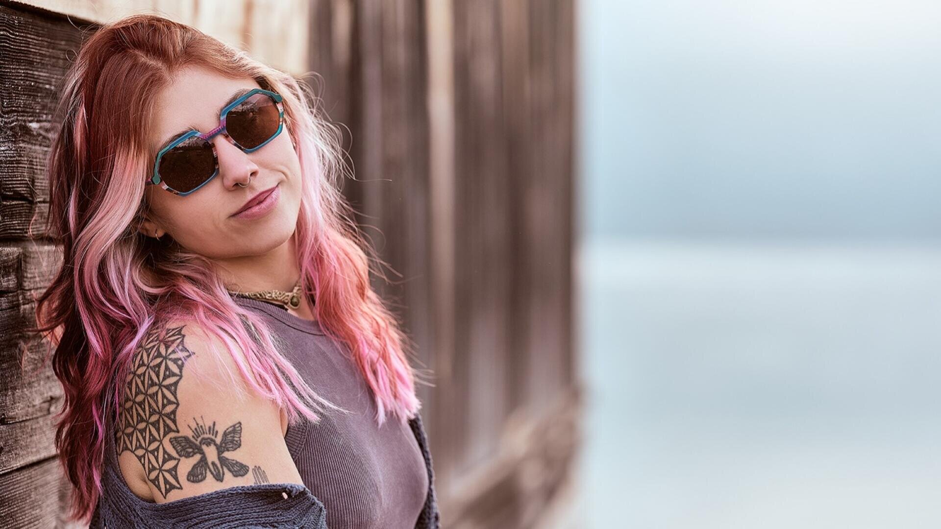 A woman with pink and blonde hair, wearing sunglasses and a sleeveless top, leans against a wooden wall. She has a geometric and a butterfly tattoo on her upper arm. The background is blurred and has a calm, light blue tone.