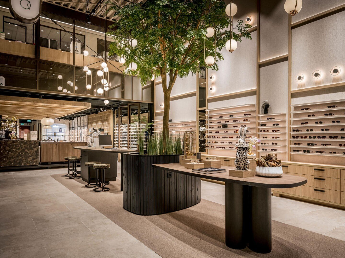 Ein modernes Brillengeschäft mit Holzregalen, auf denen Sonnenbrillen ausgestellt sind, großen dekorativen Tischen, einem zentralen Indoor-Baum, hängenden Kugelleuchten und einer stilvollen, großzügigen Einrichtung.