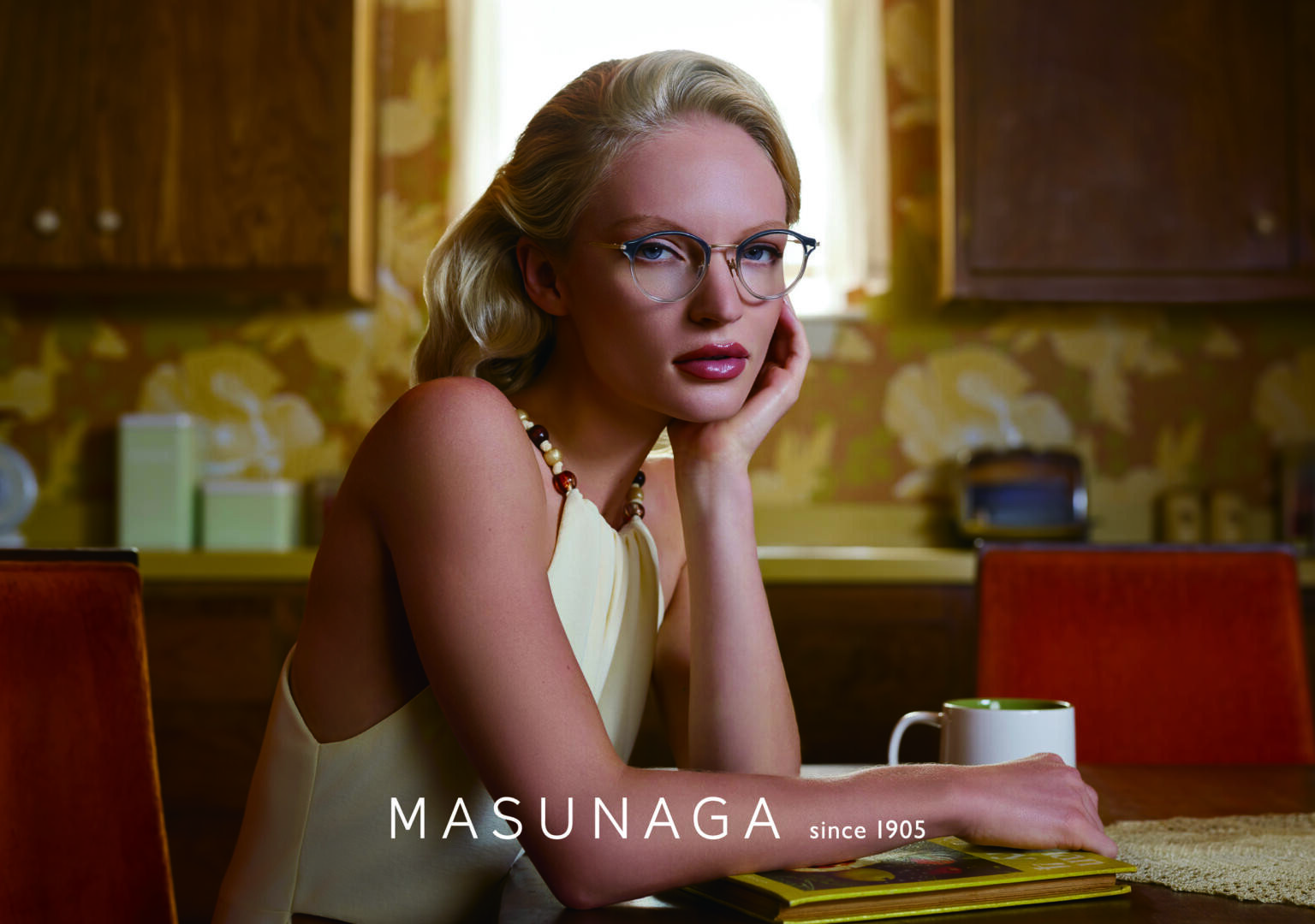 Eine Frau mit blondem Haar und Brille sitzt an einem Küchentisch mit einer weißen Tasse und einem Buch. Sie trägt ein cremefarbenes Oberteil und eine Halskette mit der Aufschrift MASUNAGA since 1905 am unteren Rand.