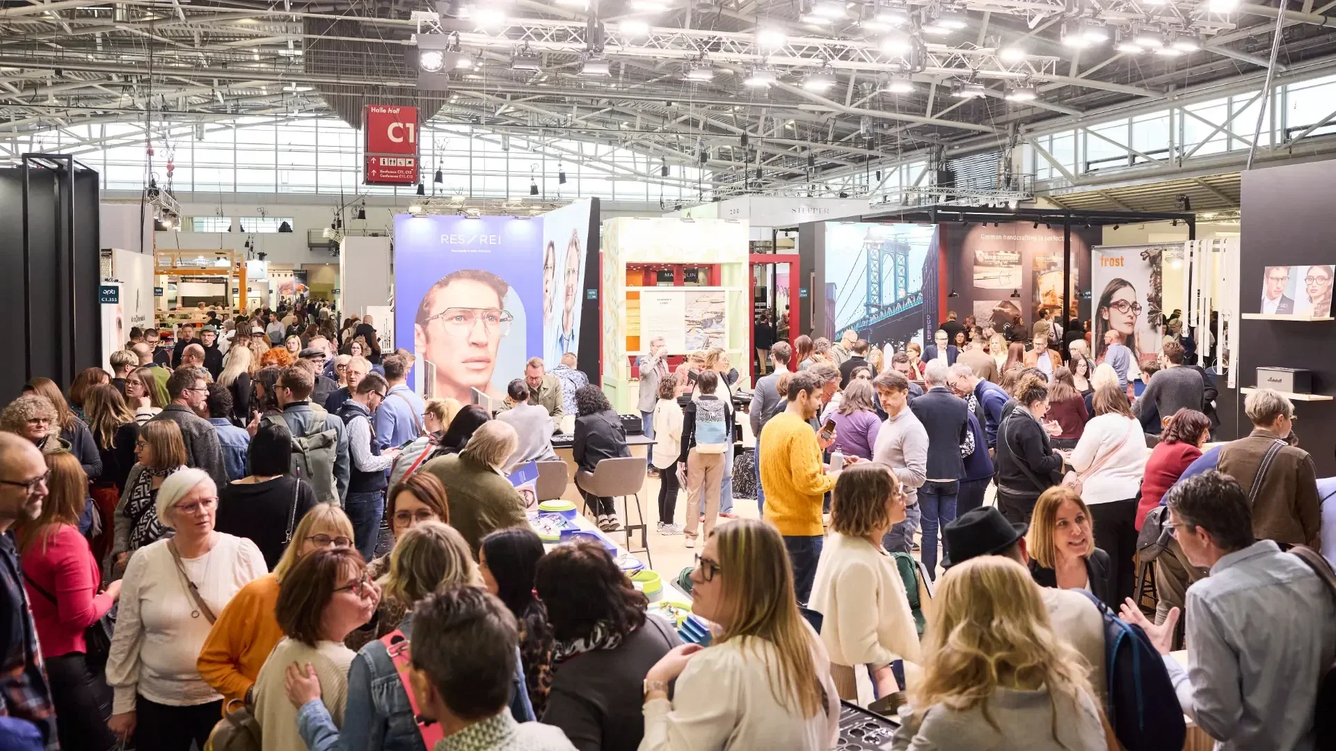 A large crowd of people gathers at an indoor convention or trade show, engaging with various booths and displays under bright lighting in a spacious hall.