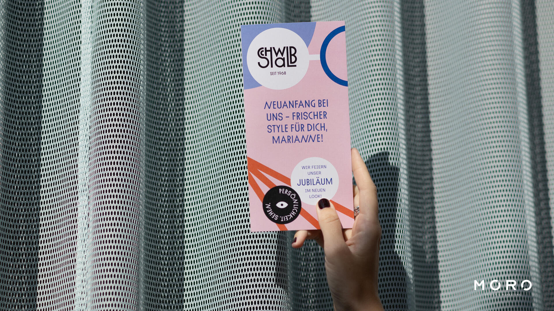 Eine Hand mit schwarzem Nagellack hält einen bunten Flyer vor einem strukturierten Vorhang. Der Flyer zeigt geometrische Formen und deutsche Texte, darunter „SCHWAB“ und „STYLE FÜR DICH, MARIANNE!“, mit dem Markenlogo MORO unten rechts.