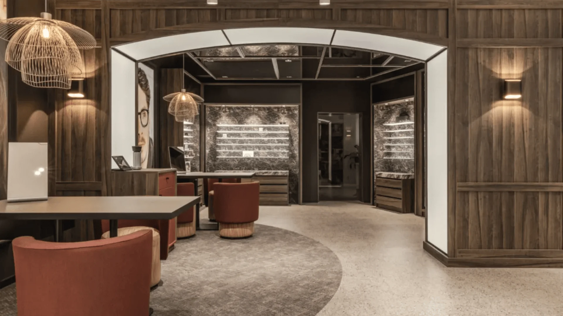 Modern, warmly lit interior with wood accents, round tables, and red upholstered chairs. Shelving with spotlights lines the back walls, and a large archway frames the stylish, inviting space.
