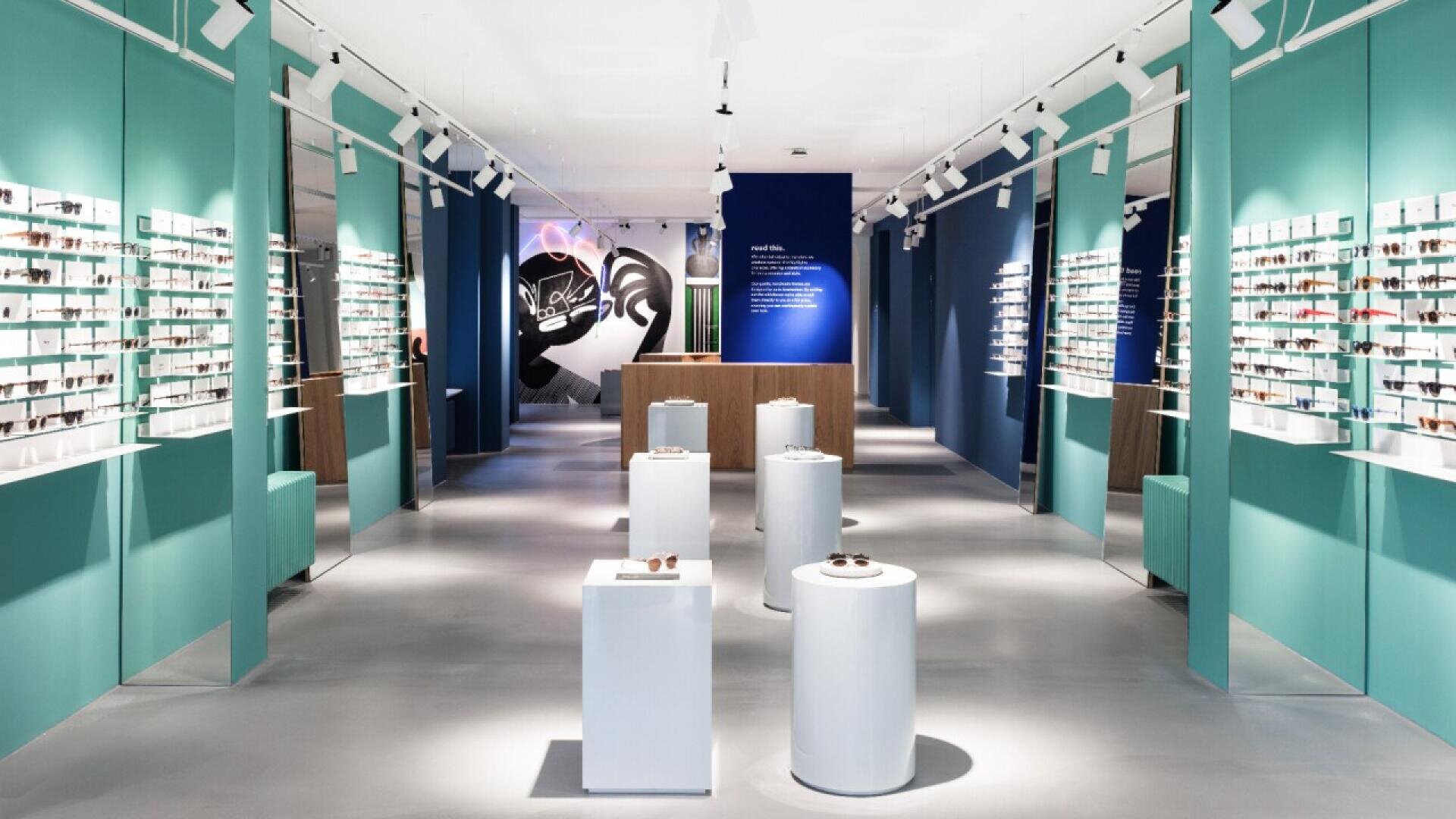 Modern eyewear store interior with teal walls, bright lighting, rows of glasses displayed on shelves, white pedestal stands showcasing sunglasses, and a wooden counter at the back.