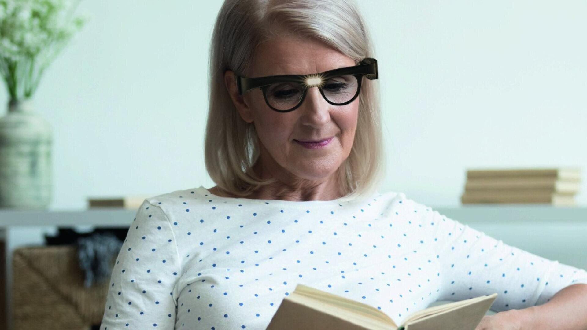An older woman with gray hair and glasses is sitting and reading a book. She is wearing a white shirt with blue polka dots, and there are books and a vase in the blurred background.