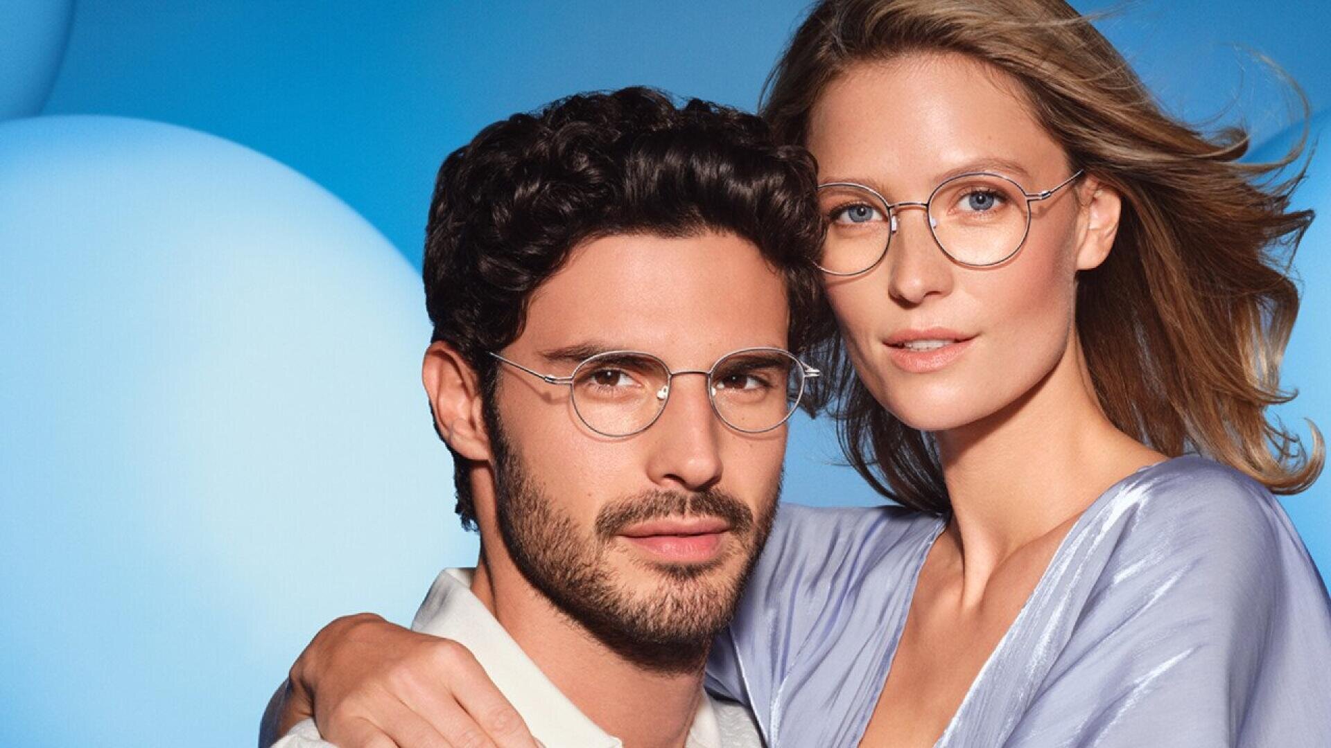 A man and a woman wearing round eyeglasses pose closely together in front of a blue background. The woman has her arm around the mans shoulder, and both have calm, neutral expressions.