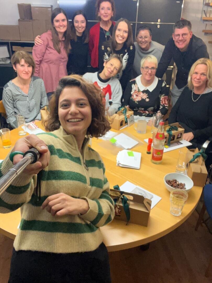 Eine Gruppe lächelnder Menschen um einen Tisch mit Getränken, Snacks und Geschenkboxen in einem gemütlichen Innenbereich macht ein Selfie. Eine Person vor ihnen hält ein Selfie-Stick, um alle auf dem Foto einzufangen.