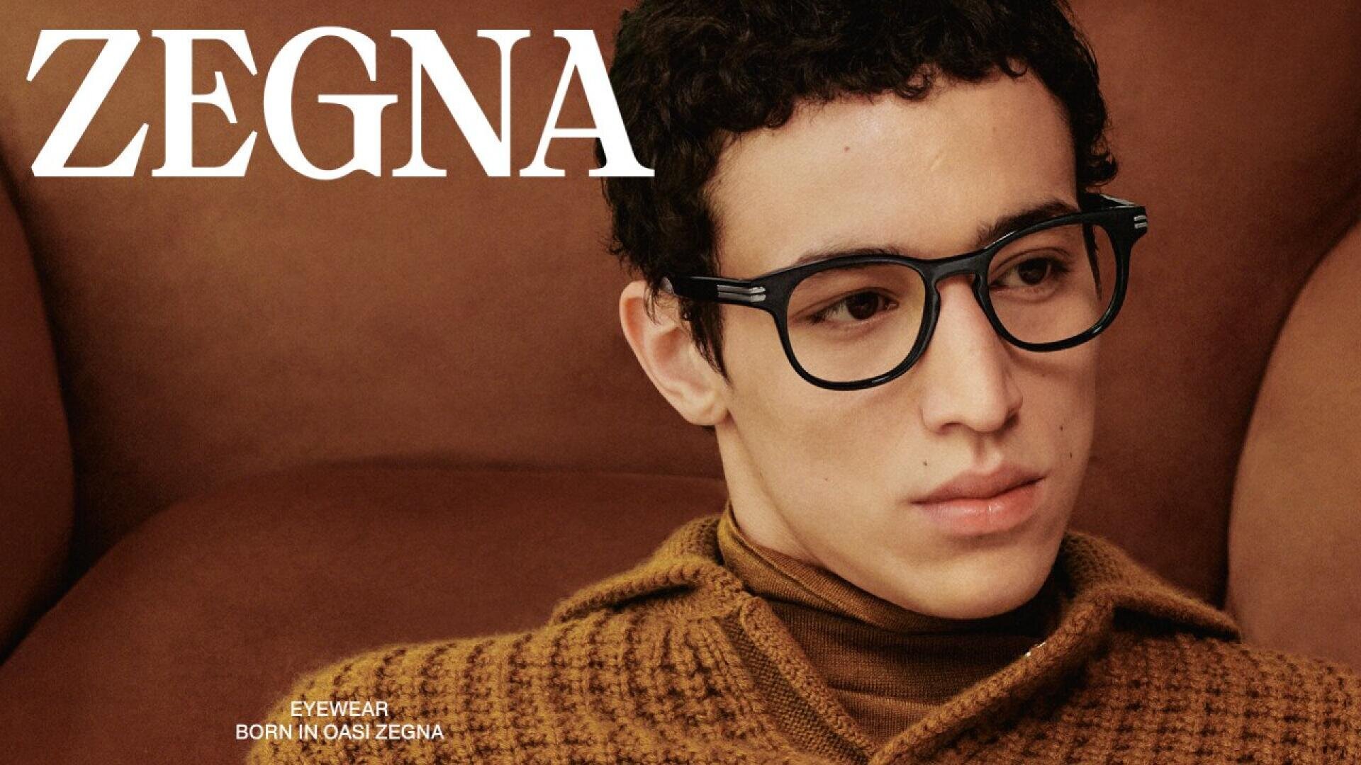 A young man wearing black glasses and a brown knit sweater sits on a brown couch. The word ZEGNA is displayed prominently in white letters at the top left, promoting the eyewear brand.