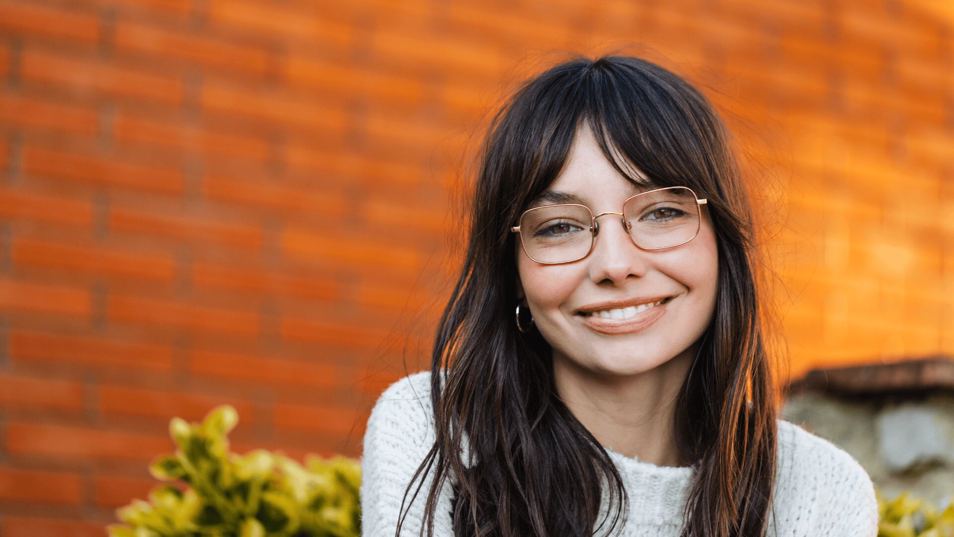 A young woman with long brown hair and glasses smiles at the camera, wearing a white sweater. An orange brick wall and green foliage are visible in the background.