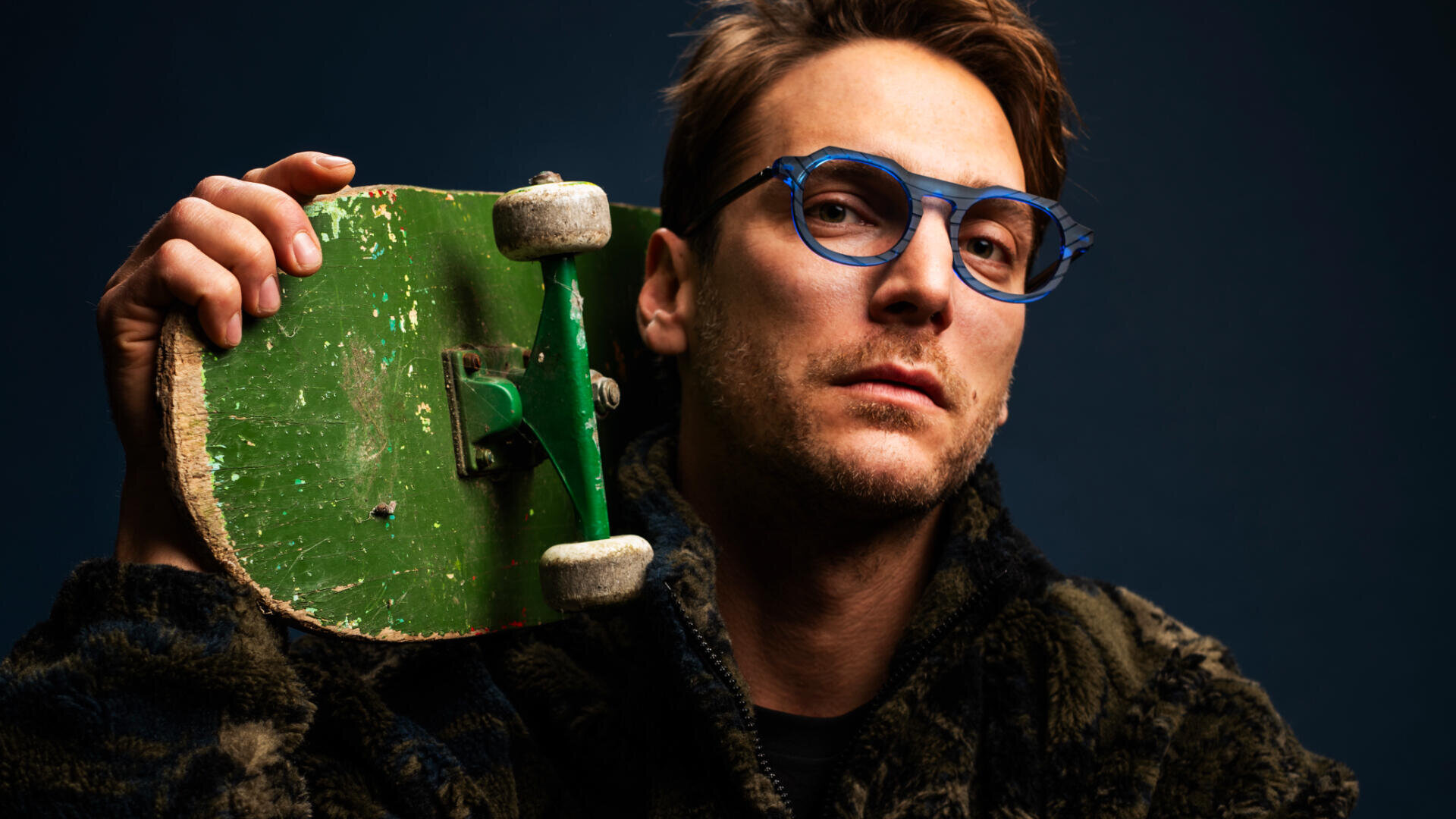 A person wearing blue glasses and a dark fuzzy jacket holds a worn green skateboard over their shoulder, looking confidently at the camera against a dark background.