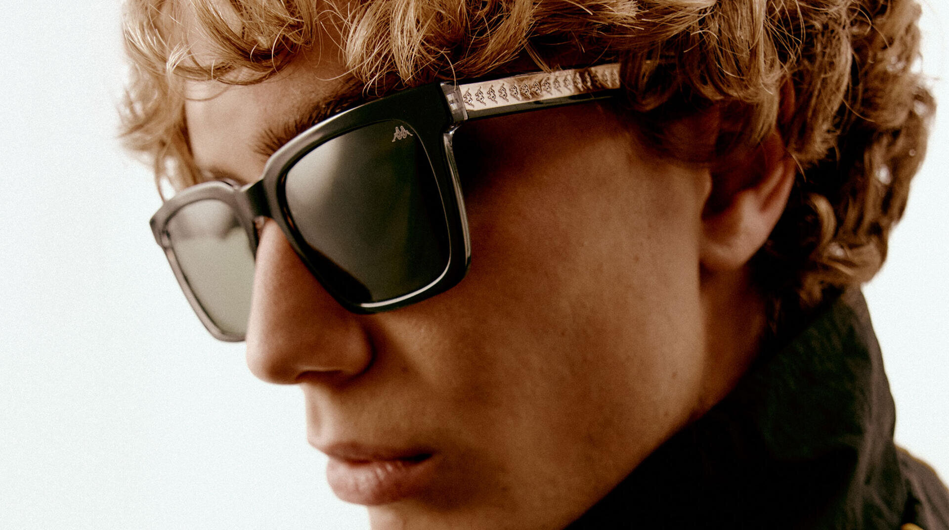 Nahaufnahme einer Person mit gewelltem blondem Haar, die eine große, quadratische schwarze Sonnenbrille und eine dunkle Jacke trägt und leicht zur Seite schaut, vor einem schlichten hellen Hintergrund.