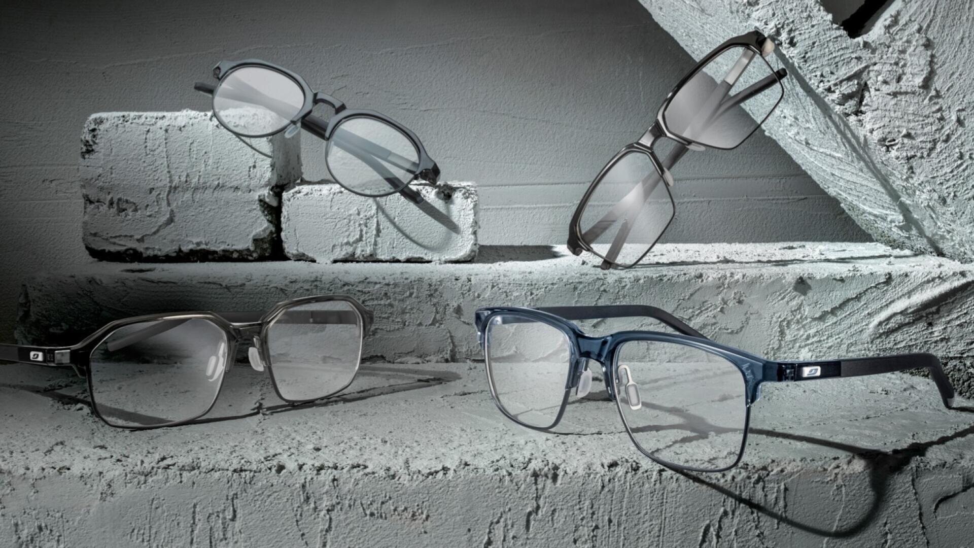 Vier Brillen mit unterschiedlichen Fassungen und Farben werden auf strukturierten, hellgrau gestrichenen Ziegeln in einer minimalistischen und künstlerischen Anordnung präsentiert.
