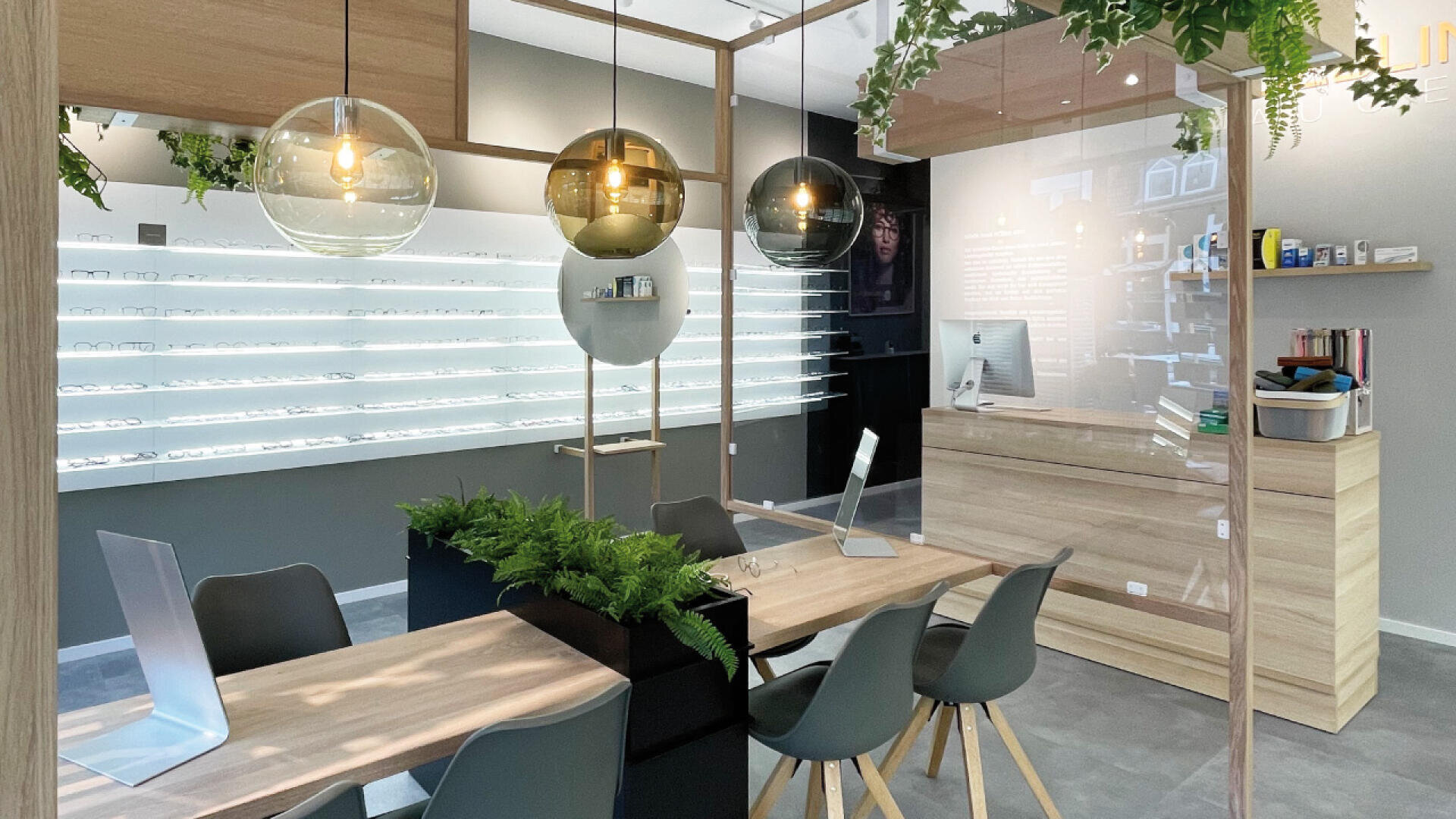 Modern optician store interior with wooden tables, black chairs, computers, and hanging pendant lights. Display shelves with eyeglasses line the wall, and a counter with computer is visible in the background. Green plants add a touch of nature.