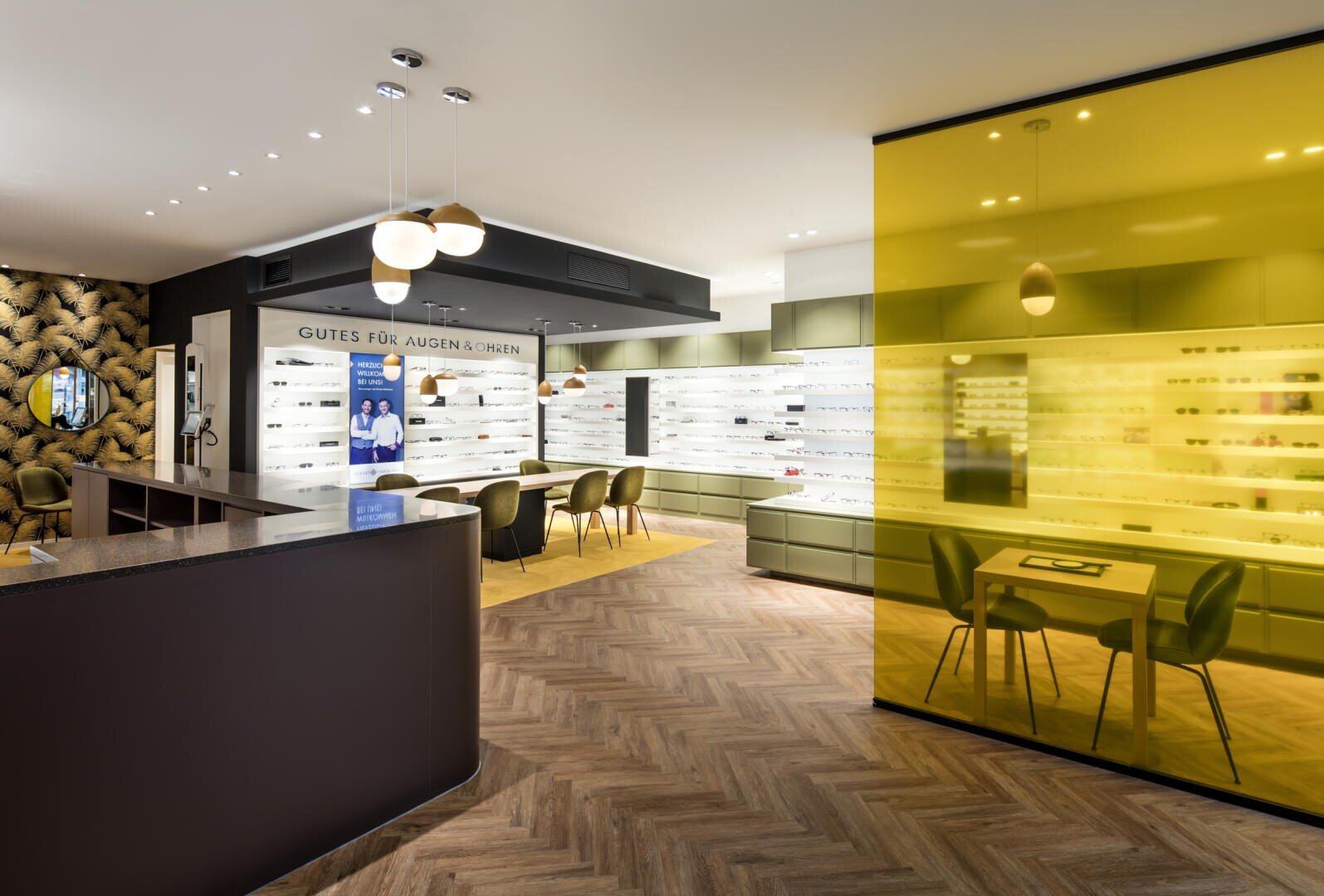 Ein modernes Optikergeschäft mit Brillenwänden, gelb getönten Glaswänden, Fischgrätenholzboden und Sitzbereichen mit schwarzen Stühlen unter Hängeleuchten.