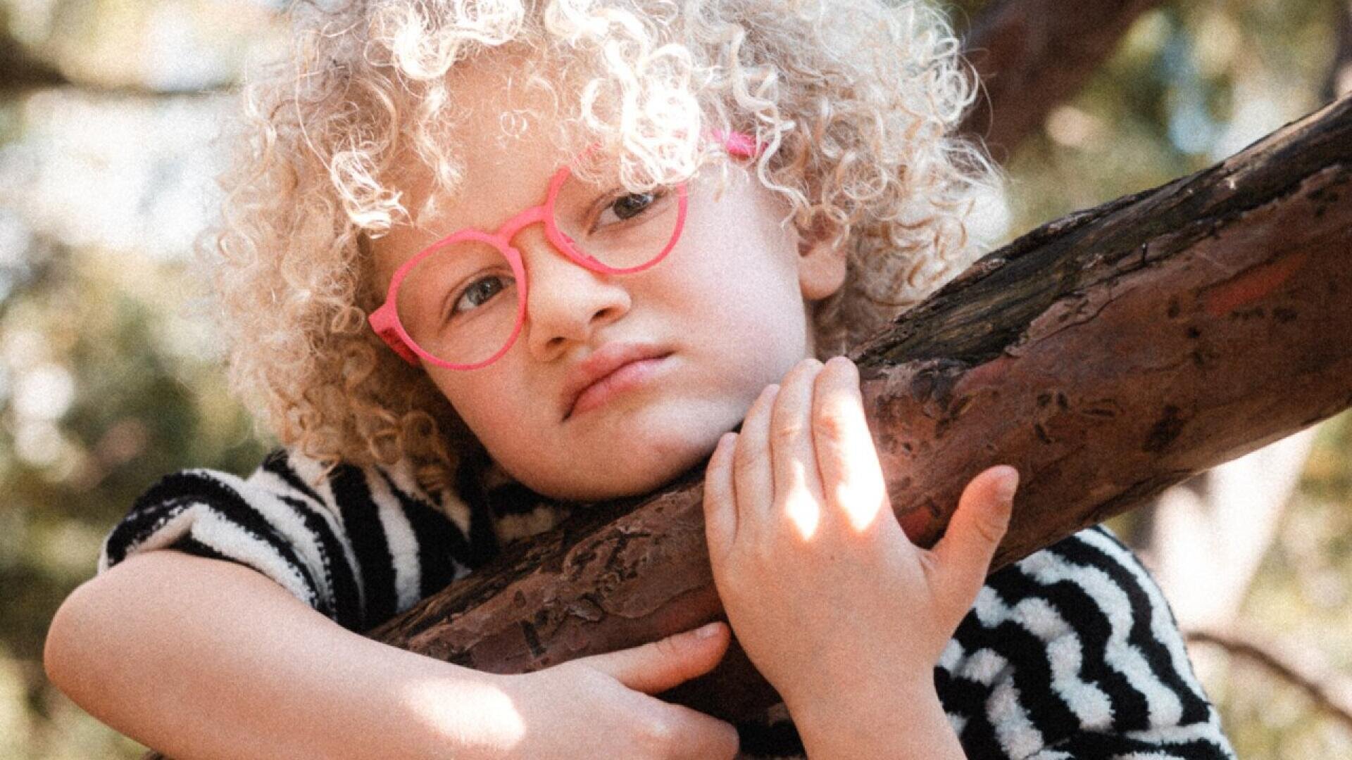 A young child with curly blond hair and pink glasses rests their arms and chin on a tree branch, looking thoughtful. The child is wearing a black and white striped top, with a blurred nature background.