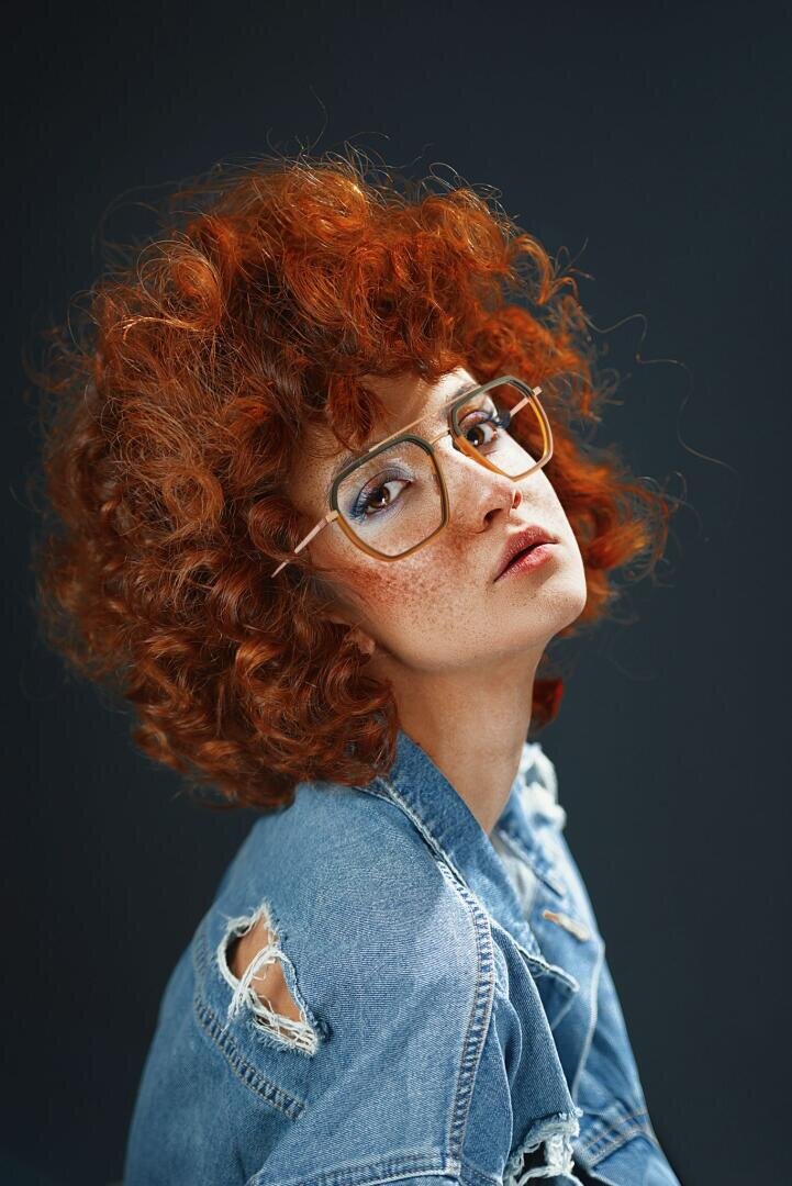 Eine Person mit lockigem rotem Haar, großer Brille und Sommersprossen blickt selbstbewusst in die Kamera, während sie eine zerrissene Jeansjacke vor einem dunklen Hintergrund trägt.