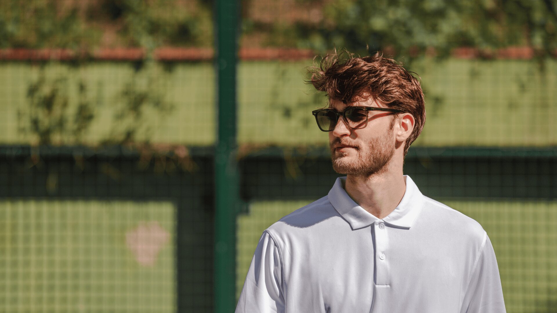 A man with short brown hair and a beard wearing dark sunglasses and a white polo shirt stands outdoors in front of a green fence.