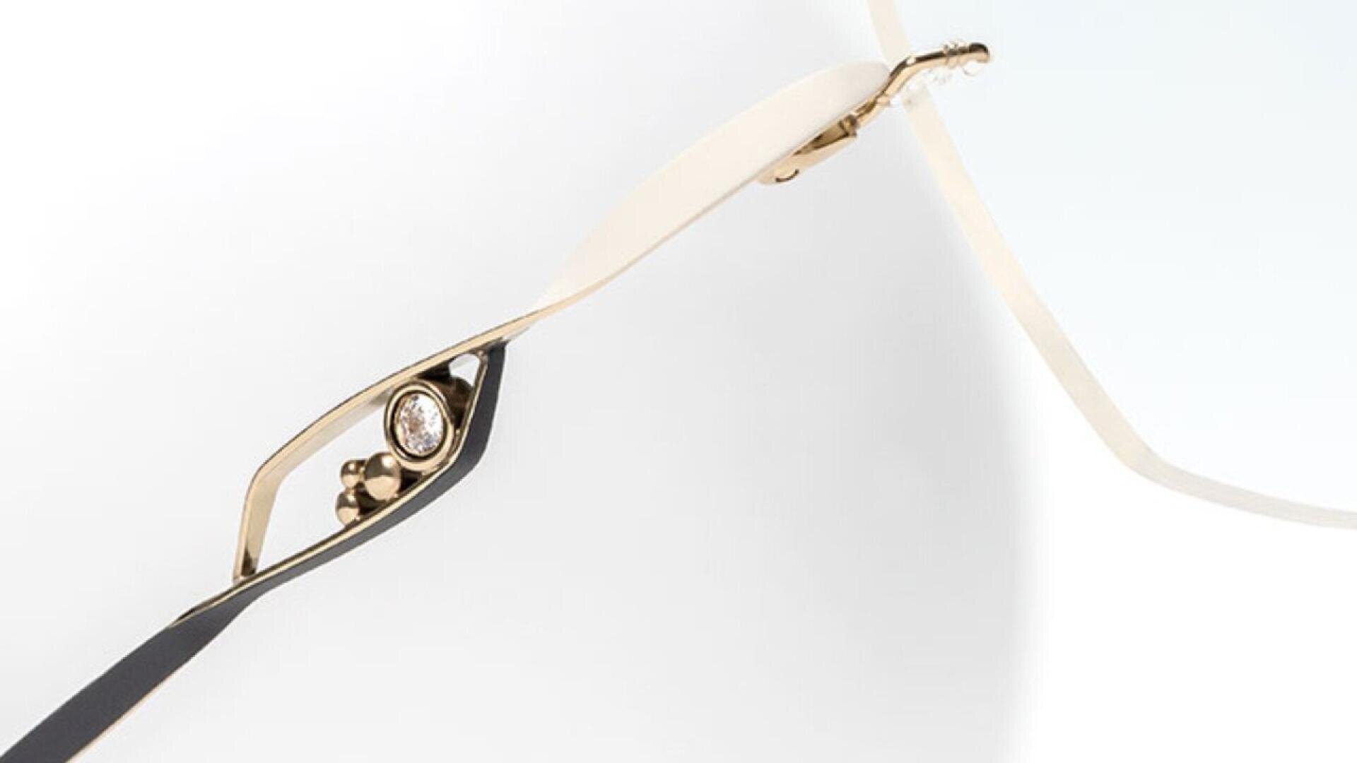 Close-up of a gold eyeglass frame with a delicate, rimless design and a small, circular gem embedded in the arm near the hinge.