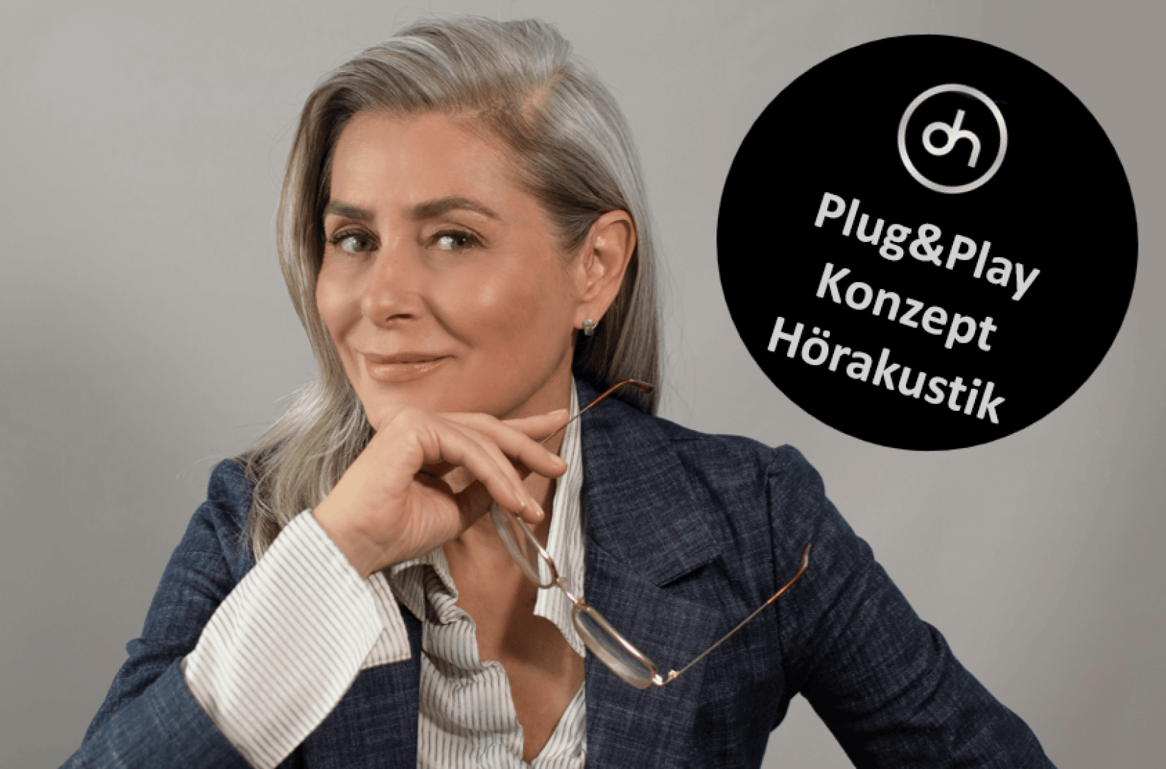 Eine selbstbewusste Frau mit grauem Haar in einem Blazer hält eine Brille und lächelt in die Kamera. Neben ihr ist ein schwarzer Kreis mit dem Text: Plug&Play Konzept Hörakustik und darüber ein Logo.