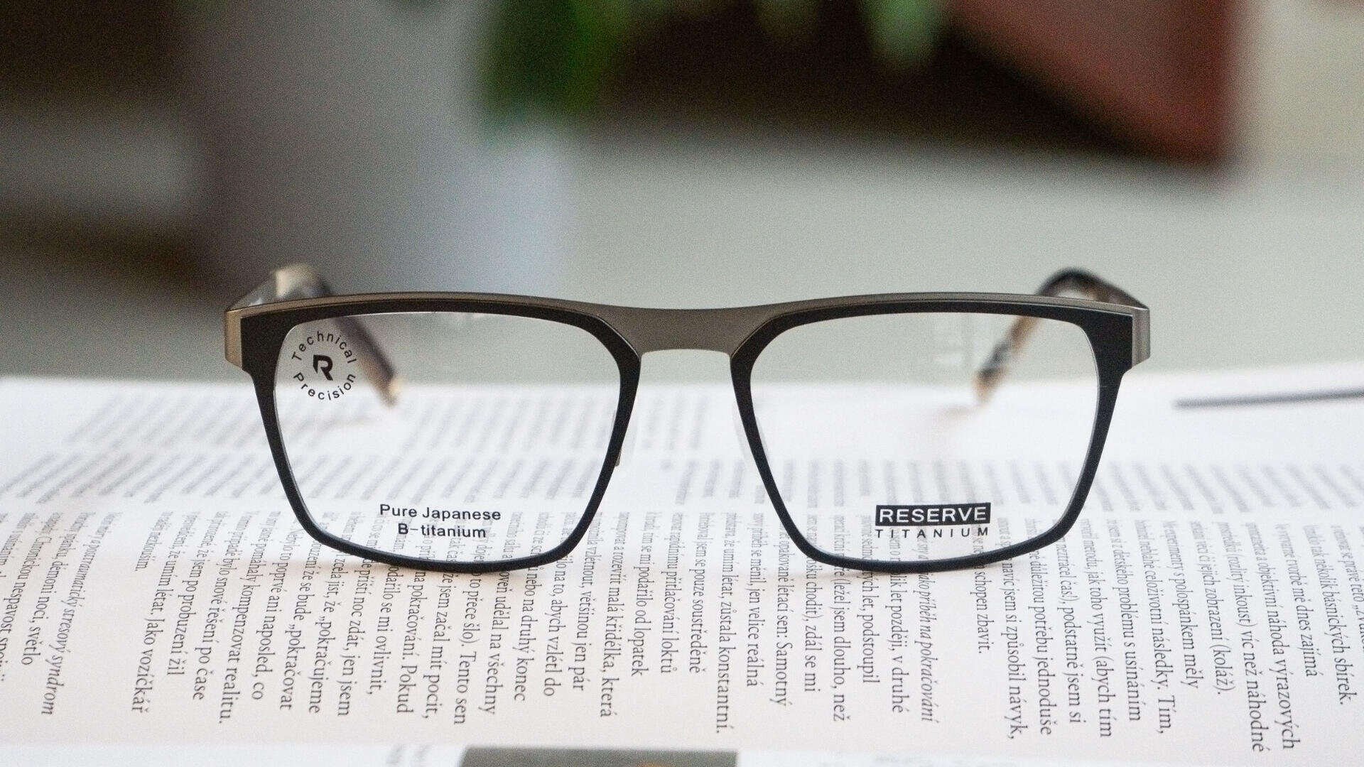 Nahaufnahme einer schwarzen rechteckigen Brille auf einem offenen Buch. Durch die Gläser ist Text sichtbar. Das linke Glas trägt den Text aus reinem japanischen β-Titan, das rechte Glas trägt den Schriftzug „RESERVE TITANIUM“.