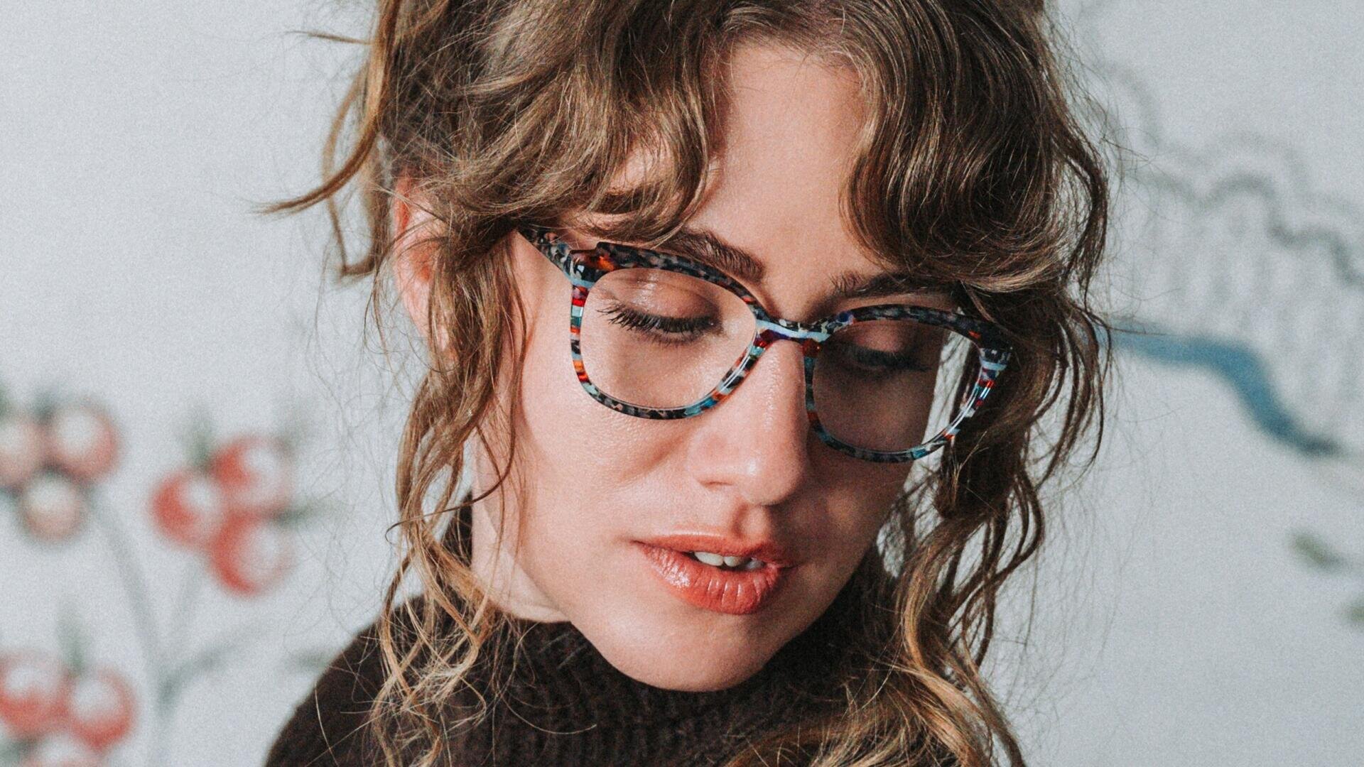 A woman with wavy light brown hair and patterned glasses looks down with a thoughtful expression. She is wearing a dark sweater and a light-colored wall with a floral pattern can be seen in the background.