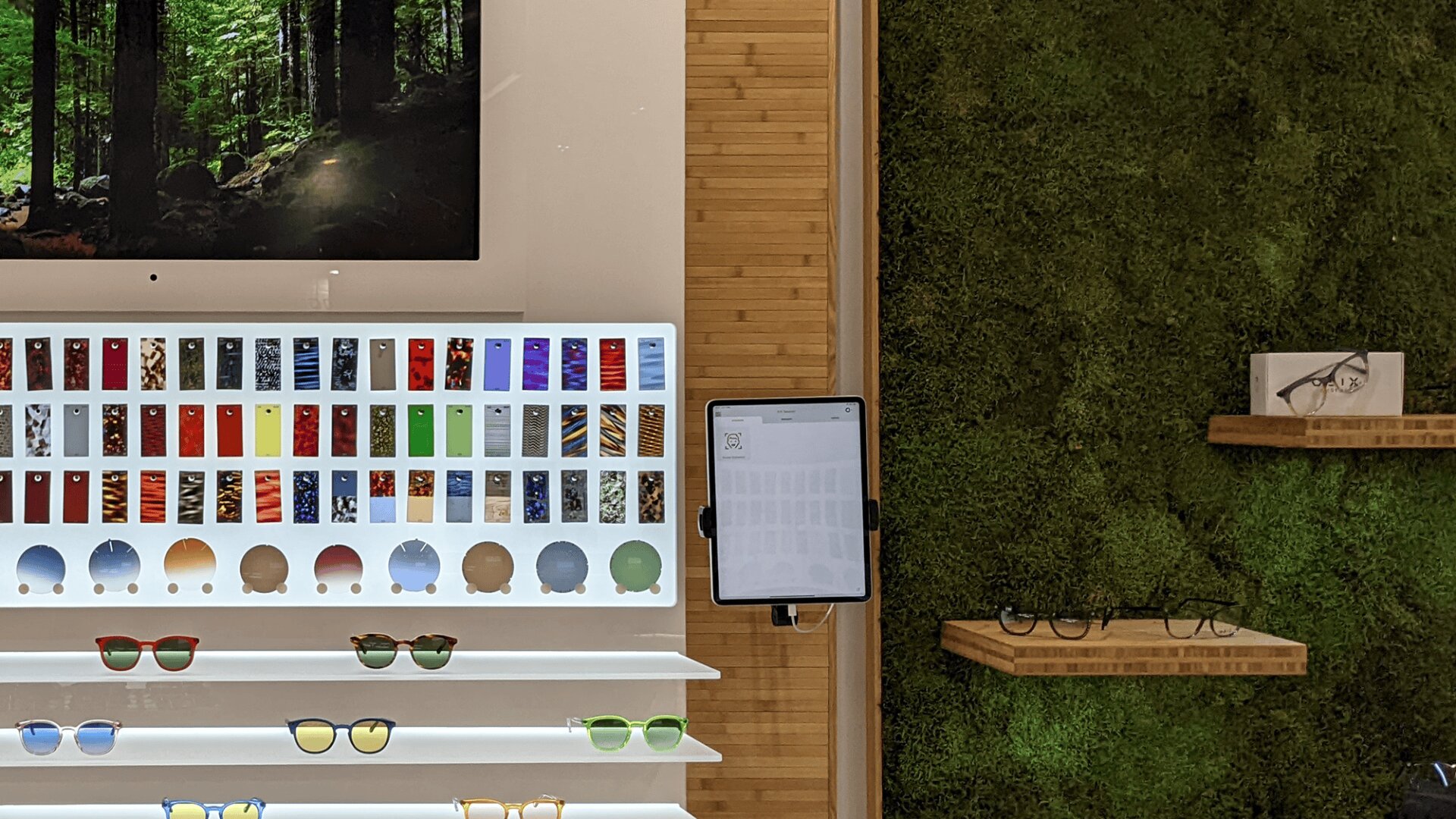 A display wall with colorful eyeglass frame samples, sunglasses on shelves, a mounted screen showing a forest, a tablet, and a green moss accent wall with eyeglasses on wooden shelves.