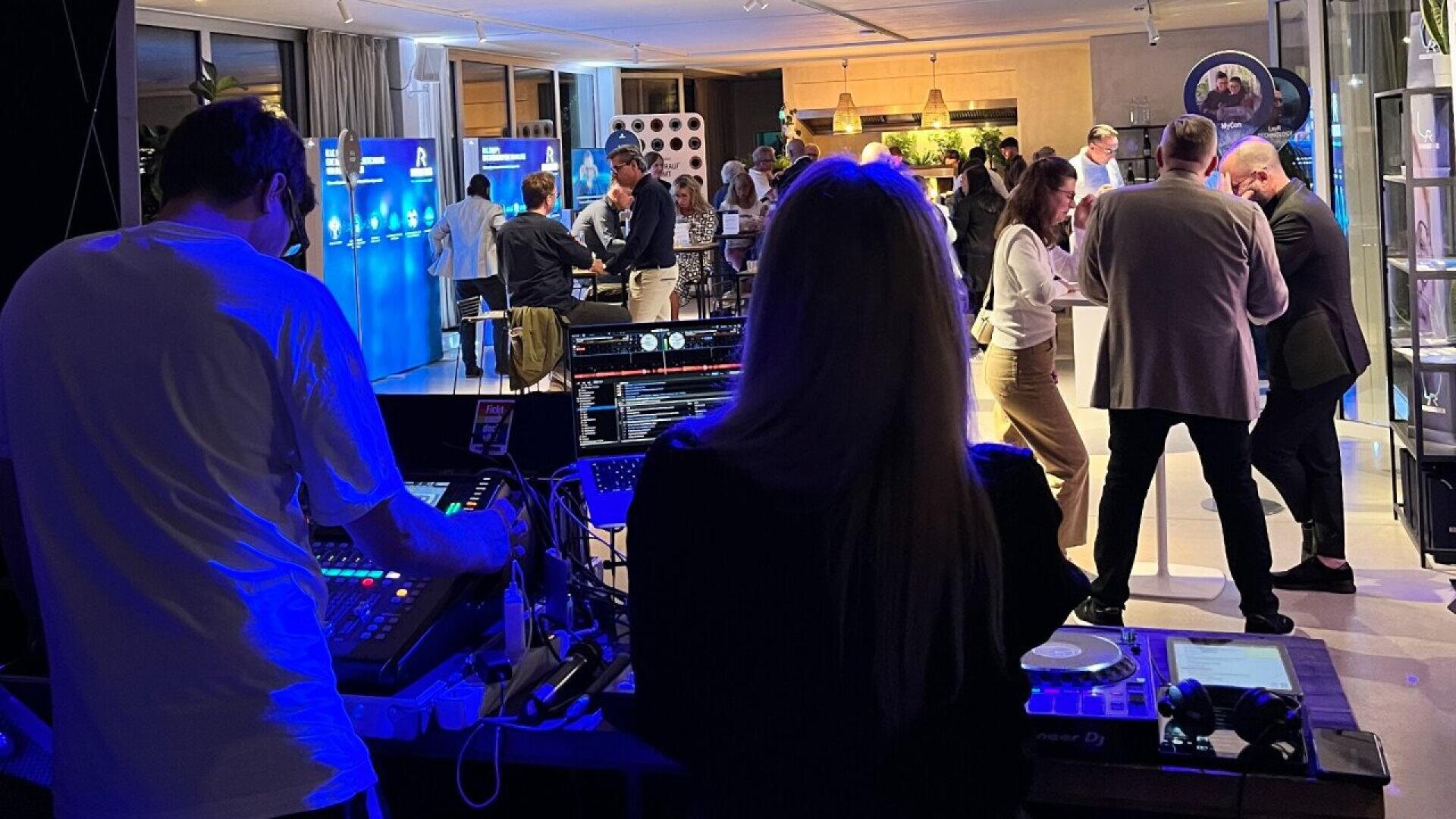 Two people manage sound and DJ equipment in the foreground at an indoor event, while groups of people are seen socializing and networking in a modern, well-lit space in the background.
