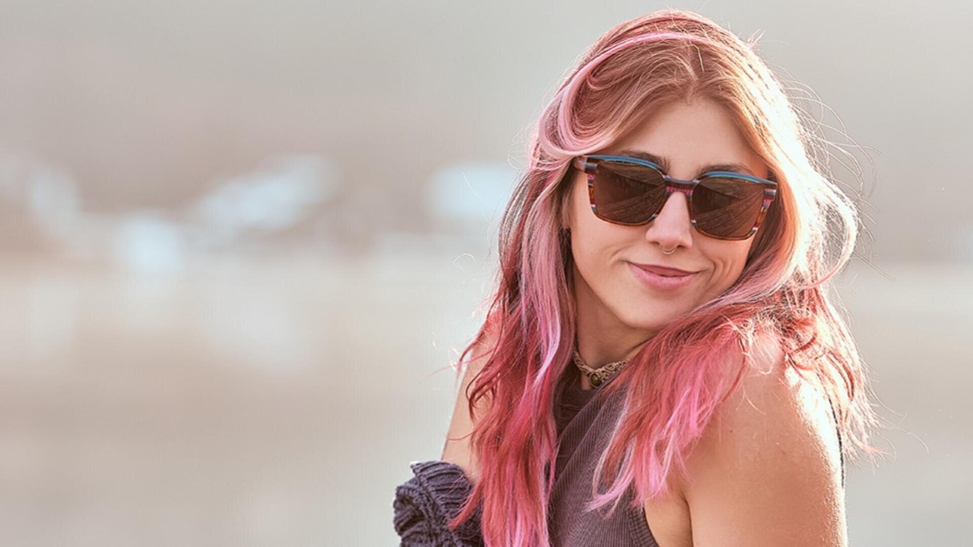 A young woman with long pink hair, sunglasses and a sleeveless top smiles slightly as she looks over her shoulder in the soft natural light outside.