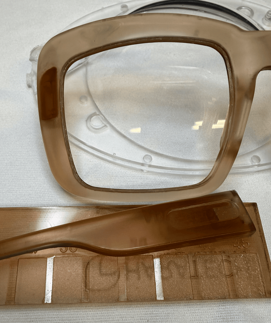 Eine Nahaufnahme eines hellbraunen Brillengestells, bei dem der Bügel auf einer Kunststoffschablone oder -form ruht, die Details des Materials und der Form zeigt.