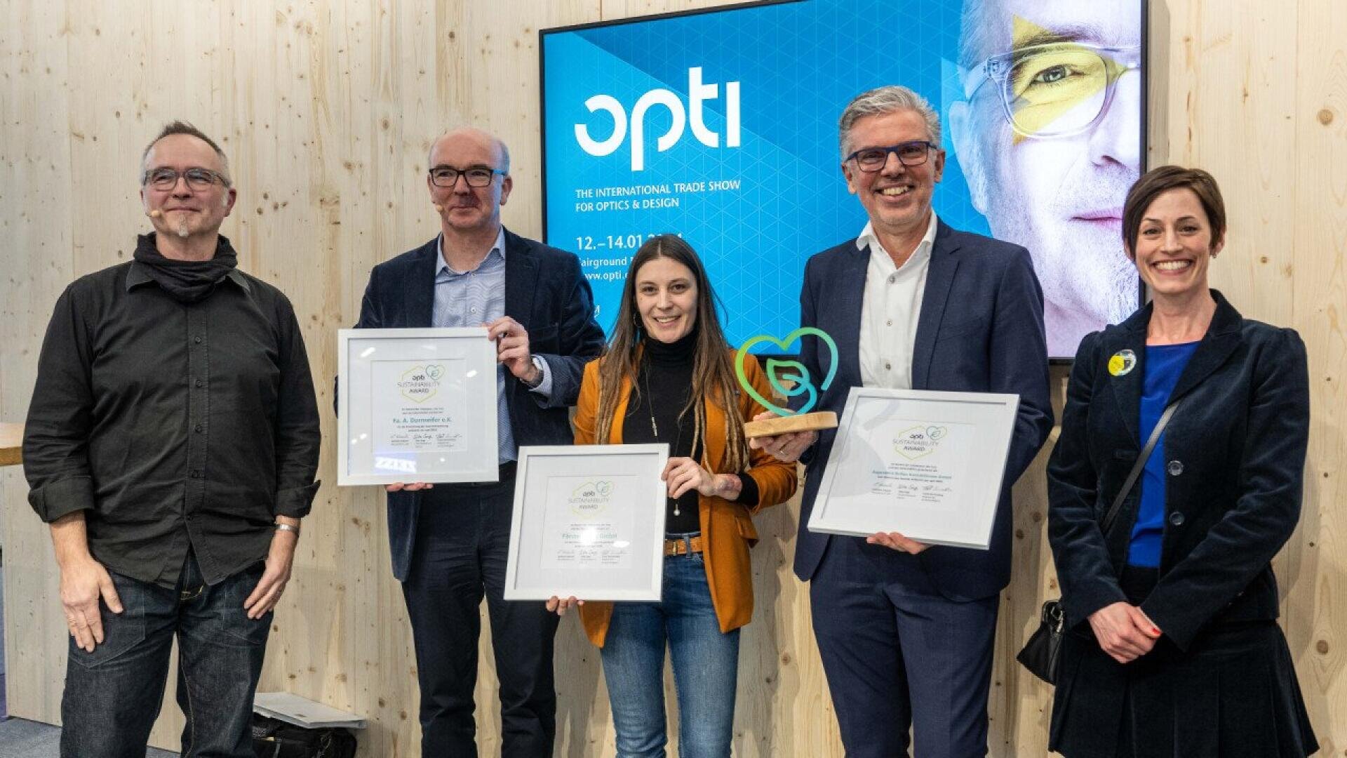 Five people stand smiling together indoors. Three hold framed certificates, and one of them also holds a green trophy. A screen behind them displays the word “opti” and event details for an optical trade show.