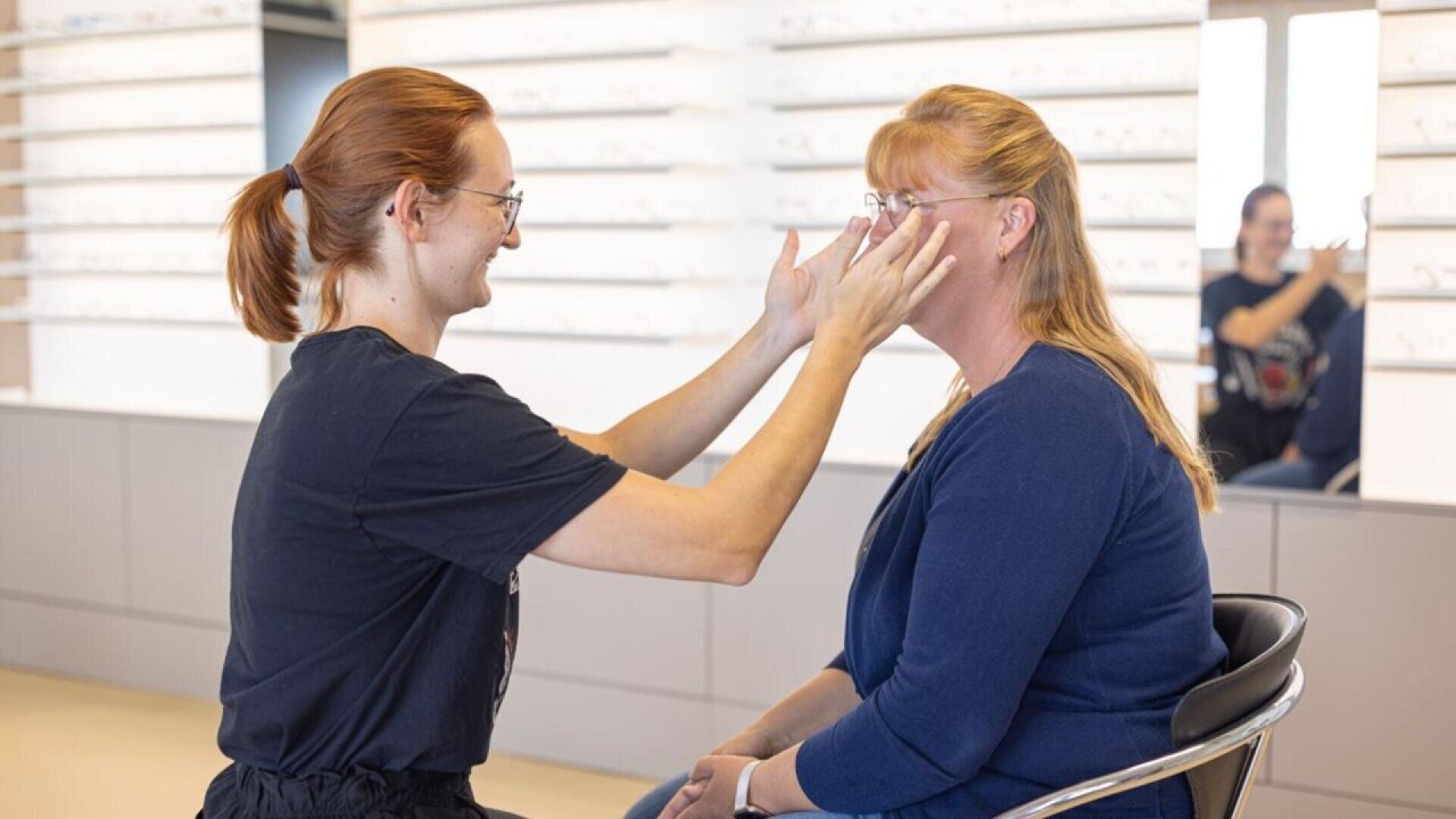 A person helps a seated woman try on eyeglasses in an optical store, both smiling. Shelves of glasses are visible in the background, and their reflection is seen in a mirror behind them.