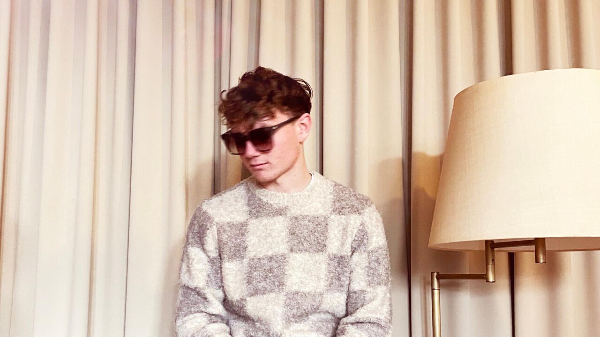 A person wearing a checkered sweater and dark sunglasses sits in front of beige curtains next to a tall floor lamp with a cream-colored lampshade.