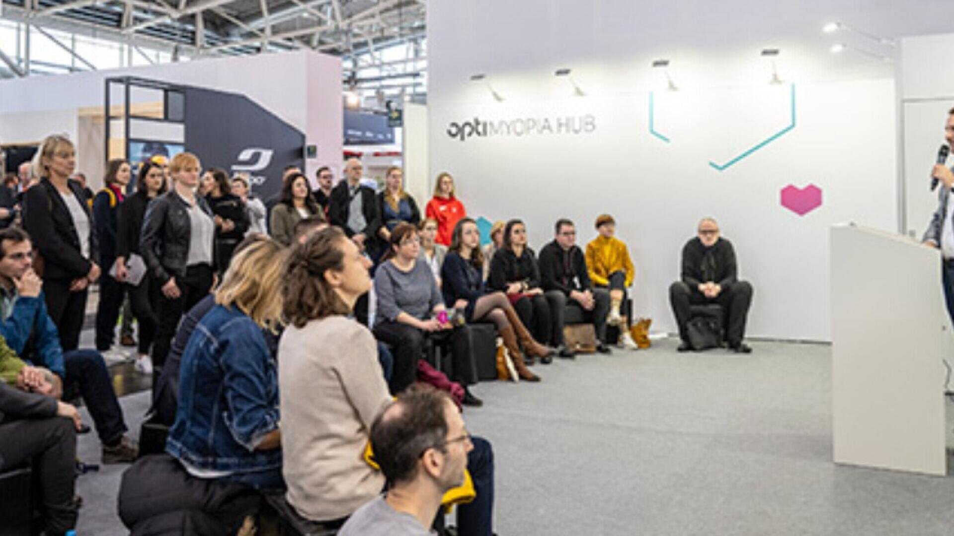 A group of people sits and stands attentively, listening to a speaker at a white podium in a modern indoor event space labeled opti FORA HUB.