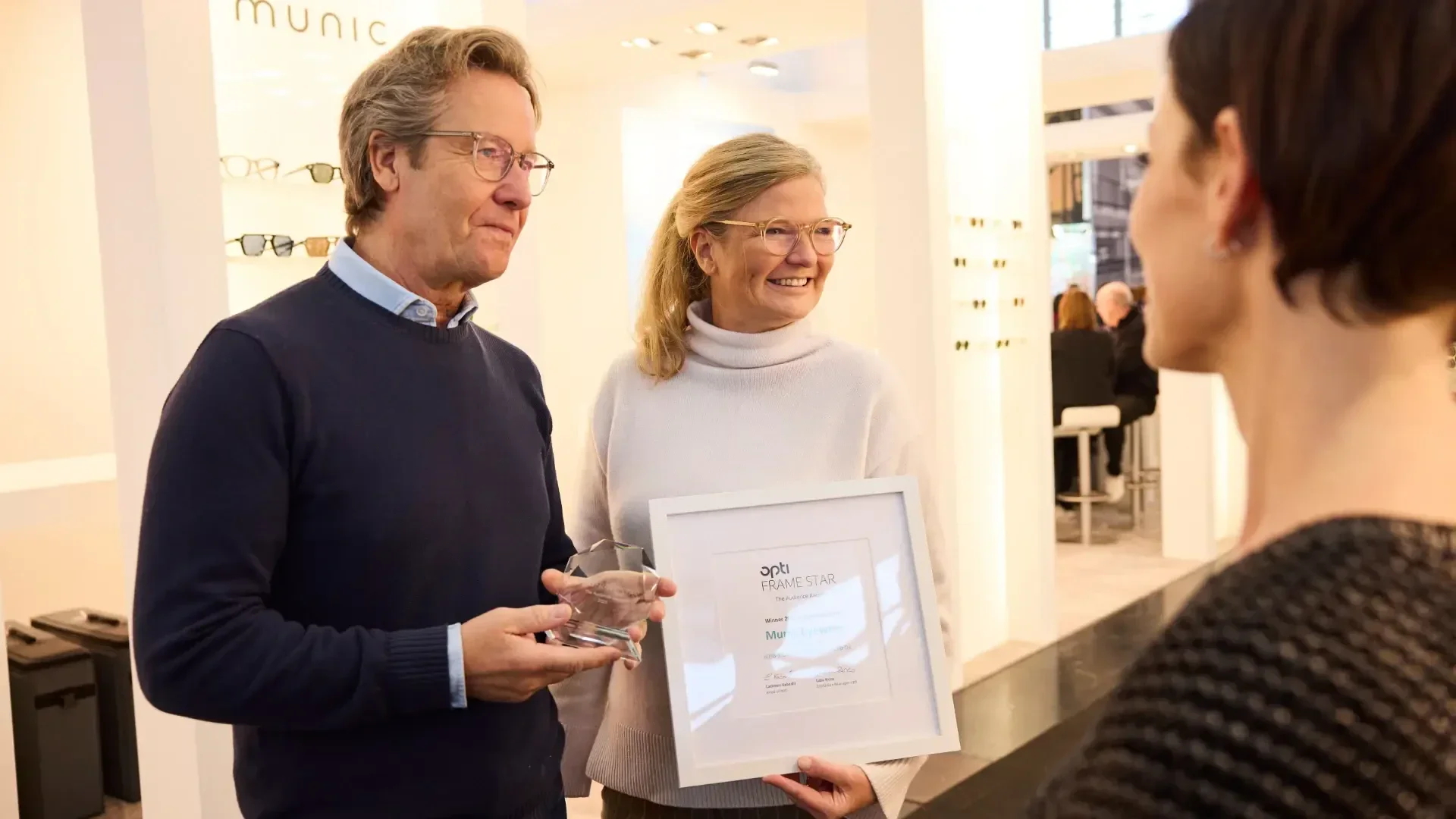Two smiling people stand indoors, one holding a glass trophy and the other a framed certificate, while facing a third person. A display of eyeglasses is visible in the background.