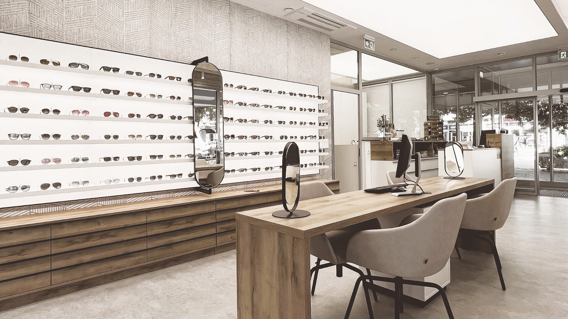 Modernes Optikergeschäft mit einer Wand mit Brillen und Sonnenbrillen, Holztischen mit Spiegeln und Stühlen sowie großen Fenstern, die natürliches Licht hereinlassen.