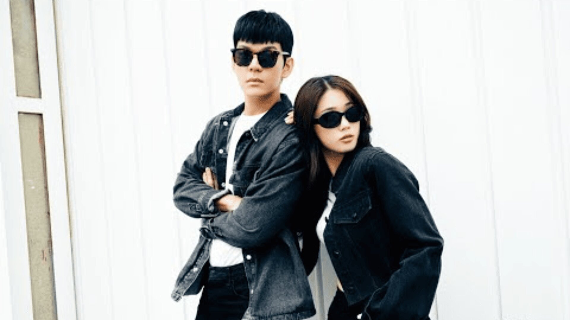 A man and woman wearing black sunglasses and black denim jackets stand confidently against a white wall, with the man crossing his arms and the woman leaning against him.