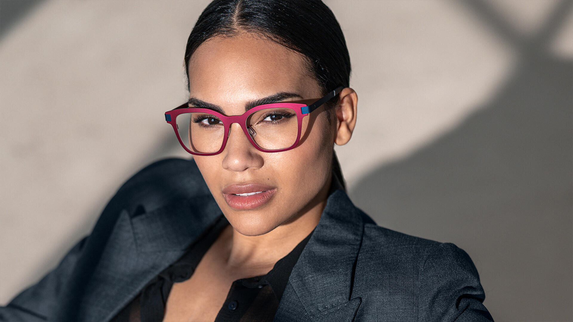 A woman with slicked-back hair wearing red-framed glasses and a dark blazer looks into the camera with a neutral expression, seated in a softly lit setting.