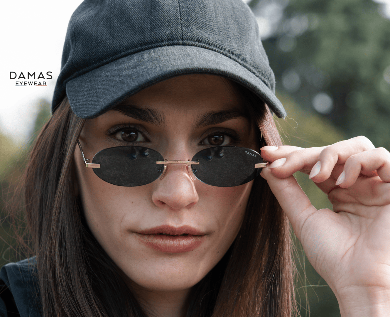 Eine Frau mit schwarzer Kappe und ovaler Sonnenbrille blickt direkt in die Kamera und hält ihre Sonnenbrille mit einer Hand. Das Logo von DAMAS EYEWEAR erscheint in der oberen linken Ecke. Die Bäume im Hintergrund sind unscharf.