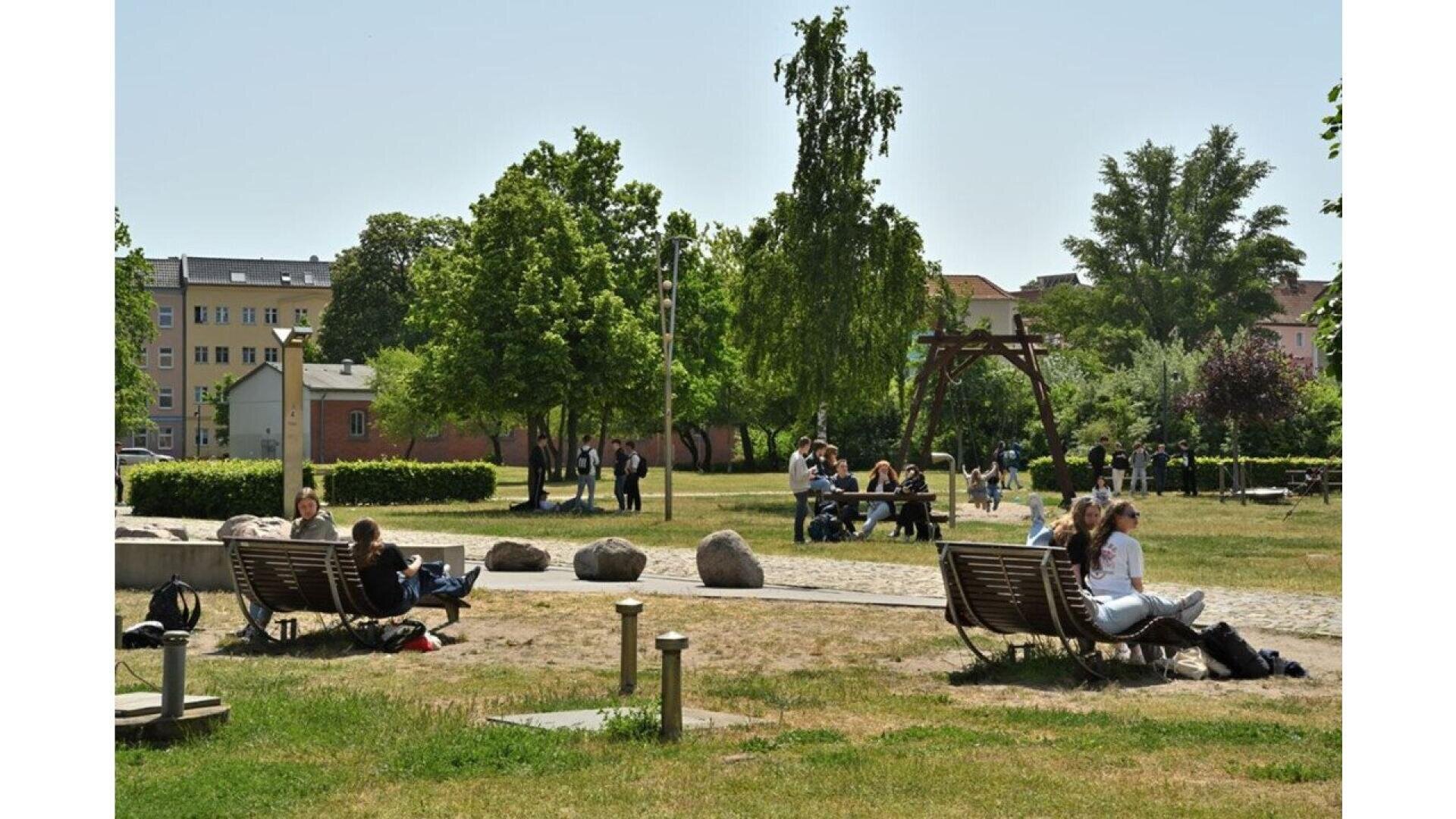 People relaxing and sitting on benches in a sunny park with green trees, grassy areas, and walking paths. Some people are talking in groups while others sit alone, with buildings visible in the background.